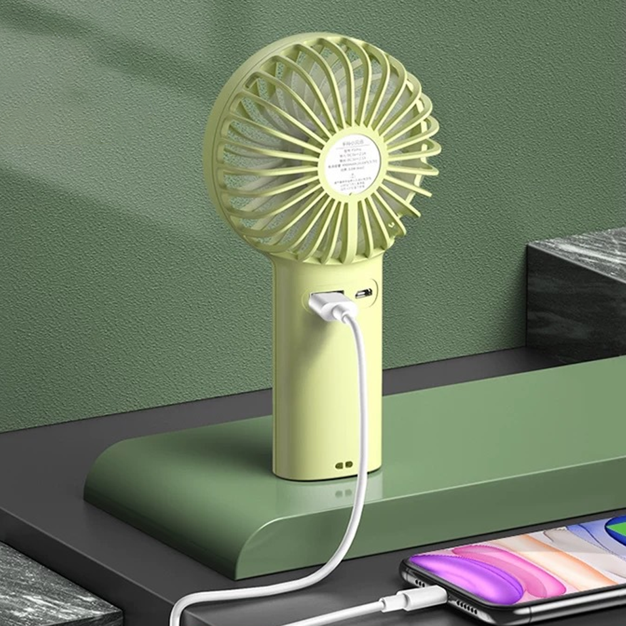 Ein grüner Handventilator wird über ein weißes USB-Kabel aufgeladen, das an ein Smartphone angeschlossen ist.