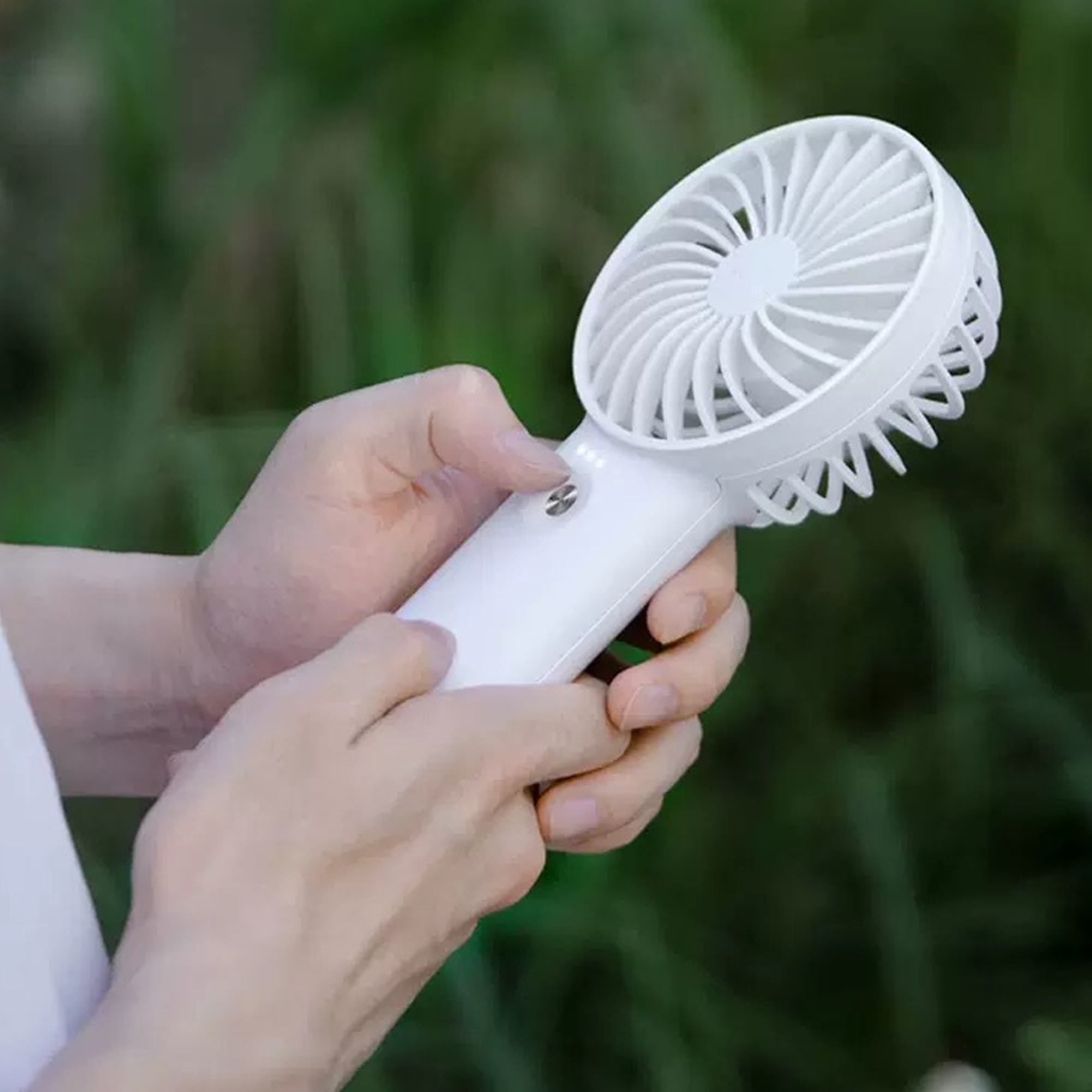 Hände halten einen weißen tragbaren Ventilator mit grünem, unscharfem Hintergrund.