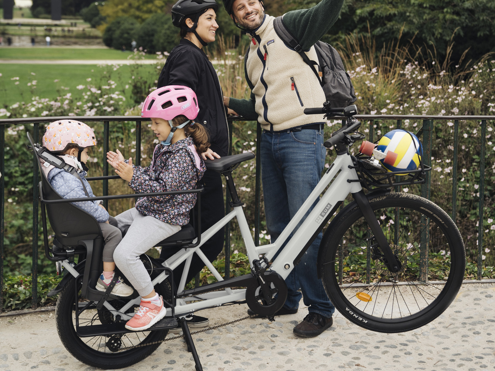 Familie mit Fahrrad im Park, Kinder auf Kindersitzen, Eltern stehen daneben.