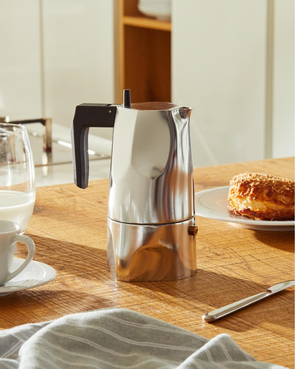 Una cafetera moka plateada sobre mesa de madera con pastel y taza de café.