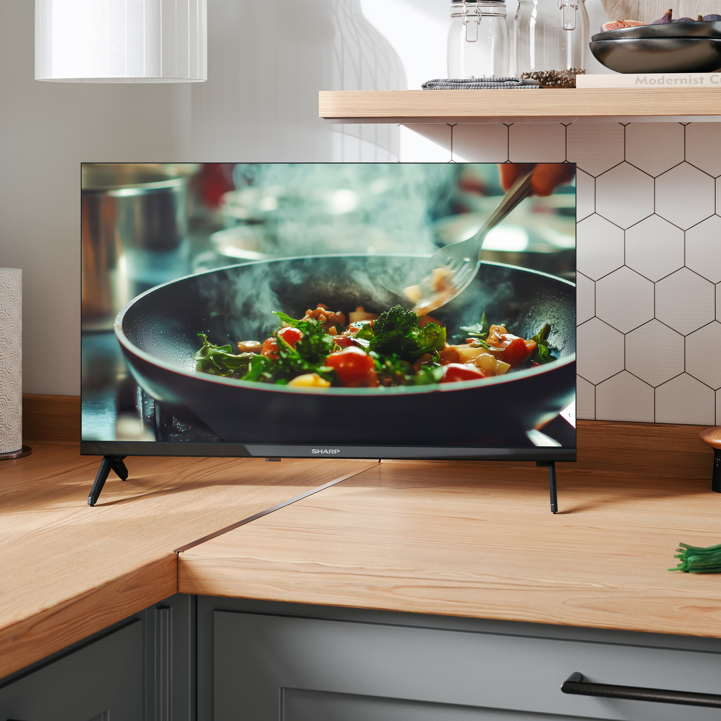 Ein Fernseher zeigt dampfendes Gemüse in einer Pfanne, auf einem Holztresen.