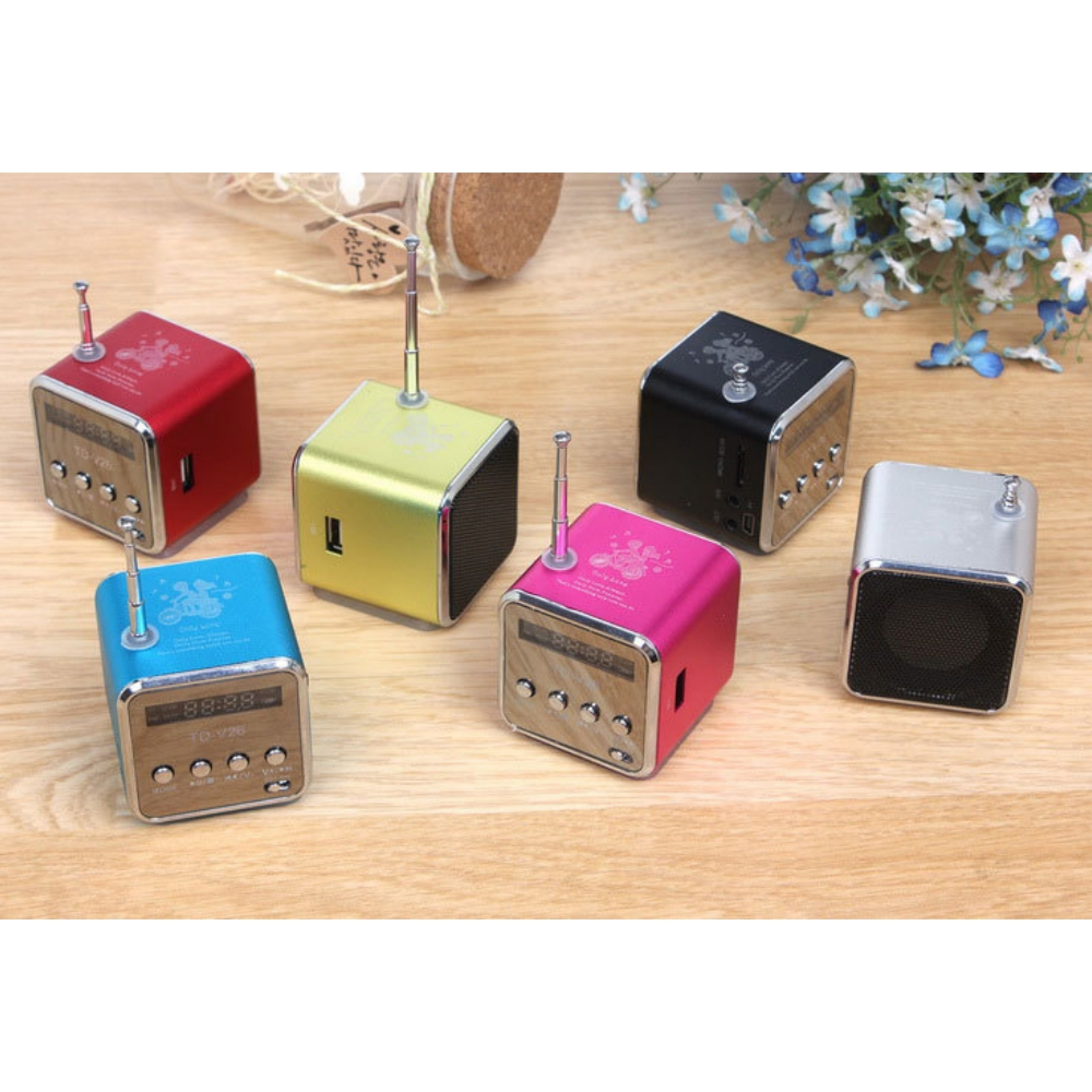 Sechs bunte Mini-Radios auf Holztisch mit Blumen und einem Glas im Hintergrund.