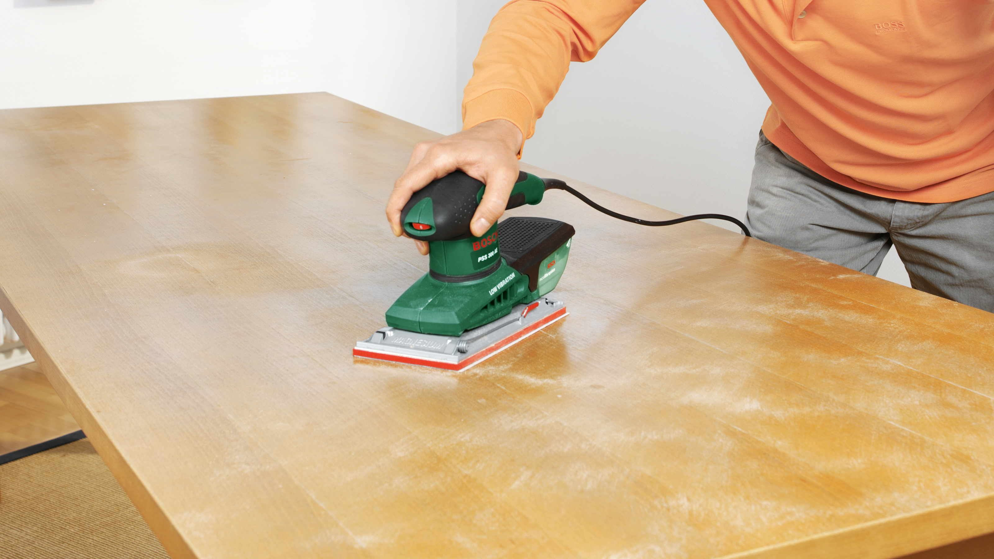 Hand schleift Holztisch mit grüner Bosch Exzenterschleifmaschine, Staub liegt auf der Oberfläche.