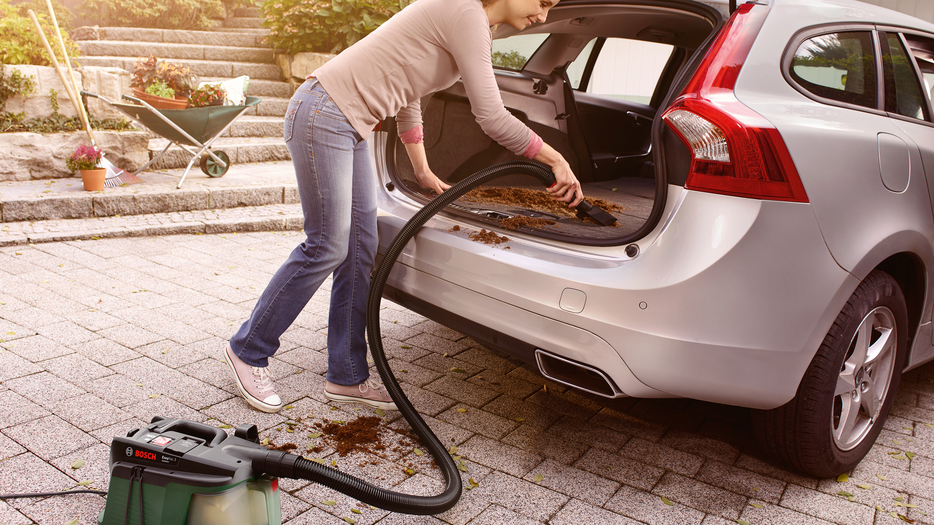 Donna aspira il bagagliaio dell'auto. Attrezzi da giardino sullo sfondo.