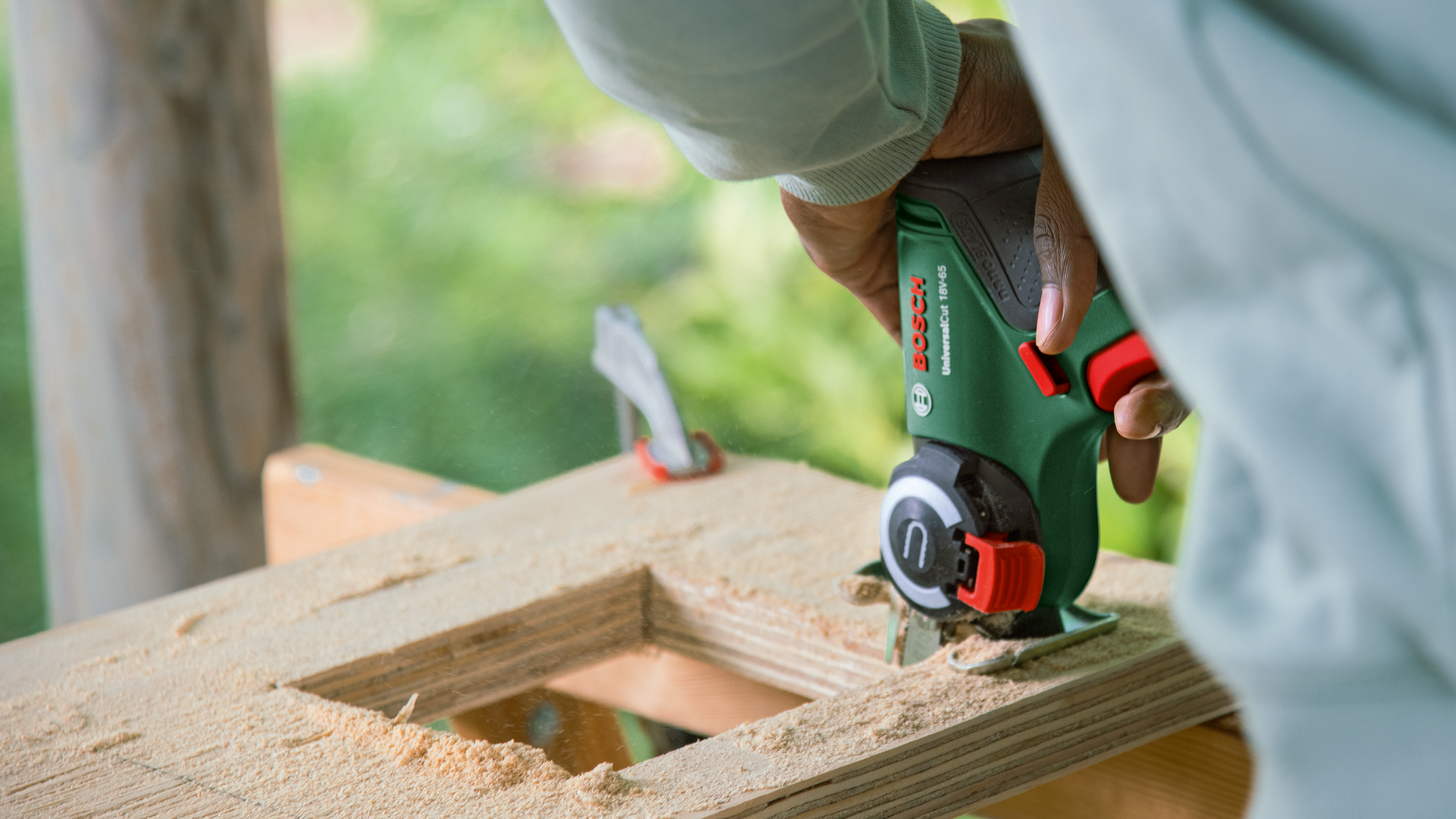 A hand holds a green Bosch jigsaw cutting wood. Sawdust is scattered around.