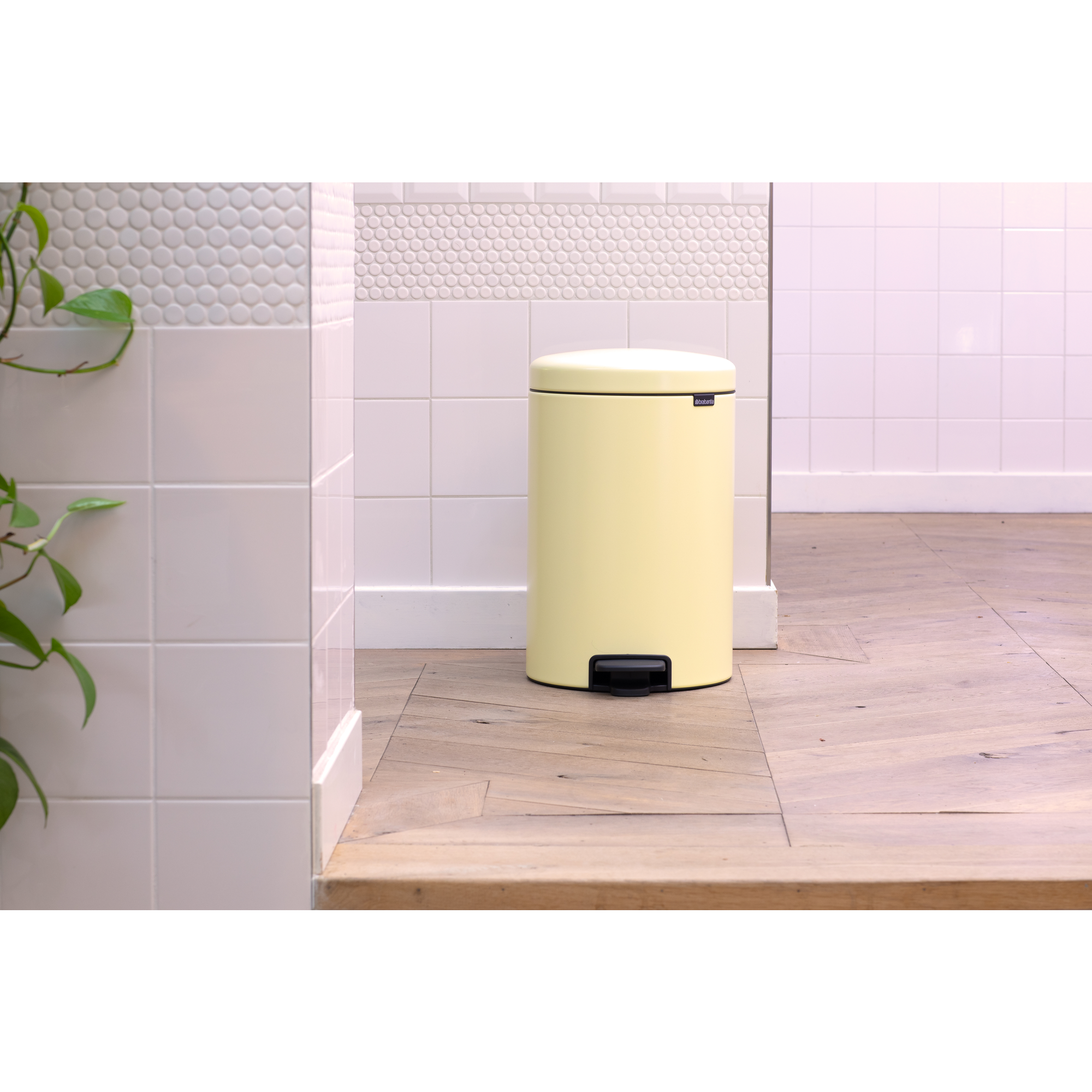 Un cubo amarillo pálido con pedal negro está sobre suelo de madera junto a azulejos blancos.