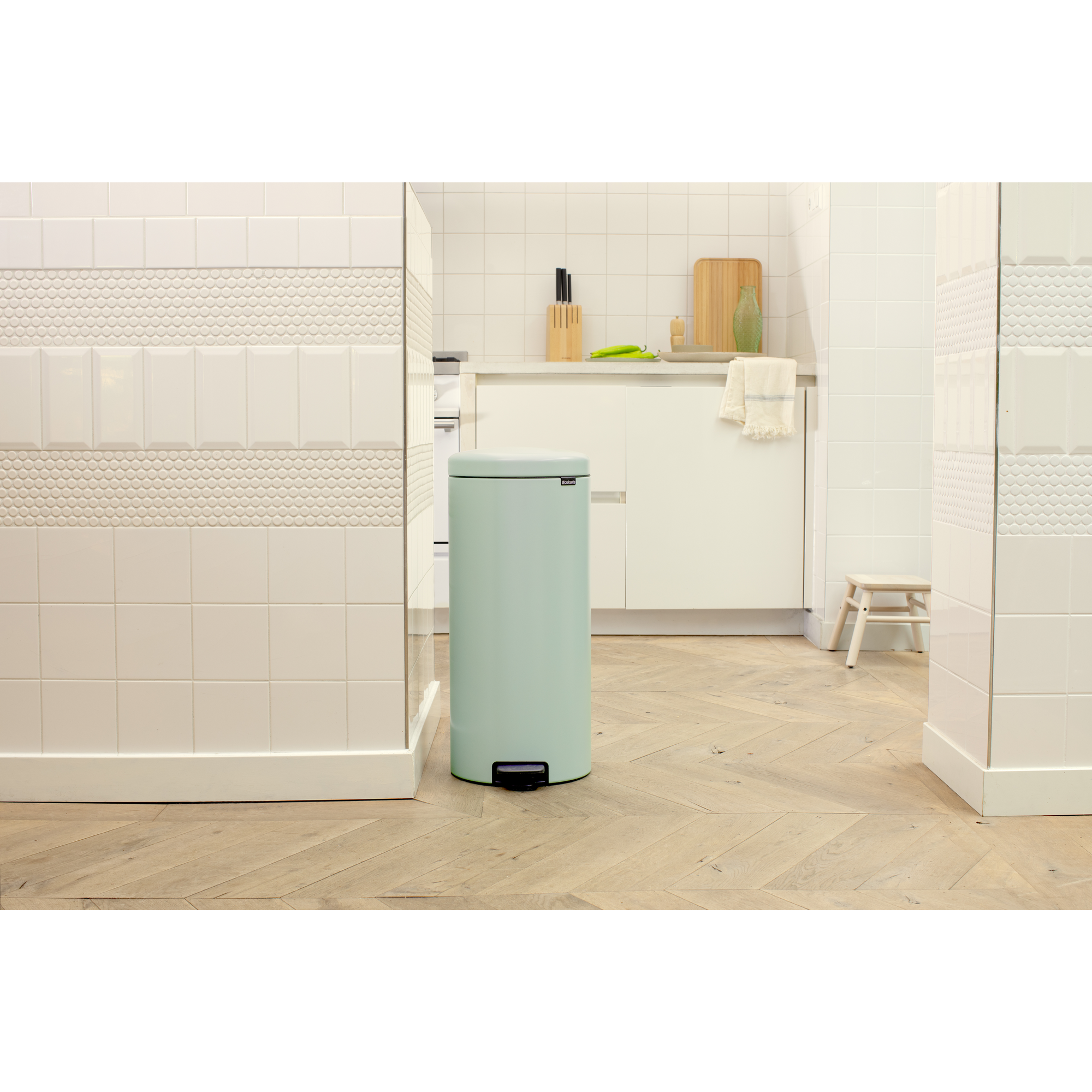Un cubo de basura de pedal verde pálido sobre suelo de madera en cocina con azulejos blancos.
