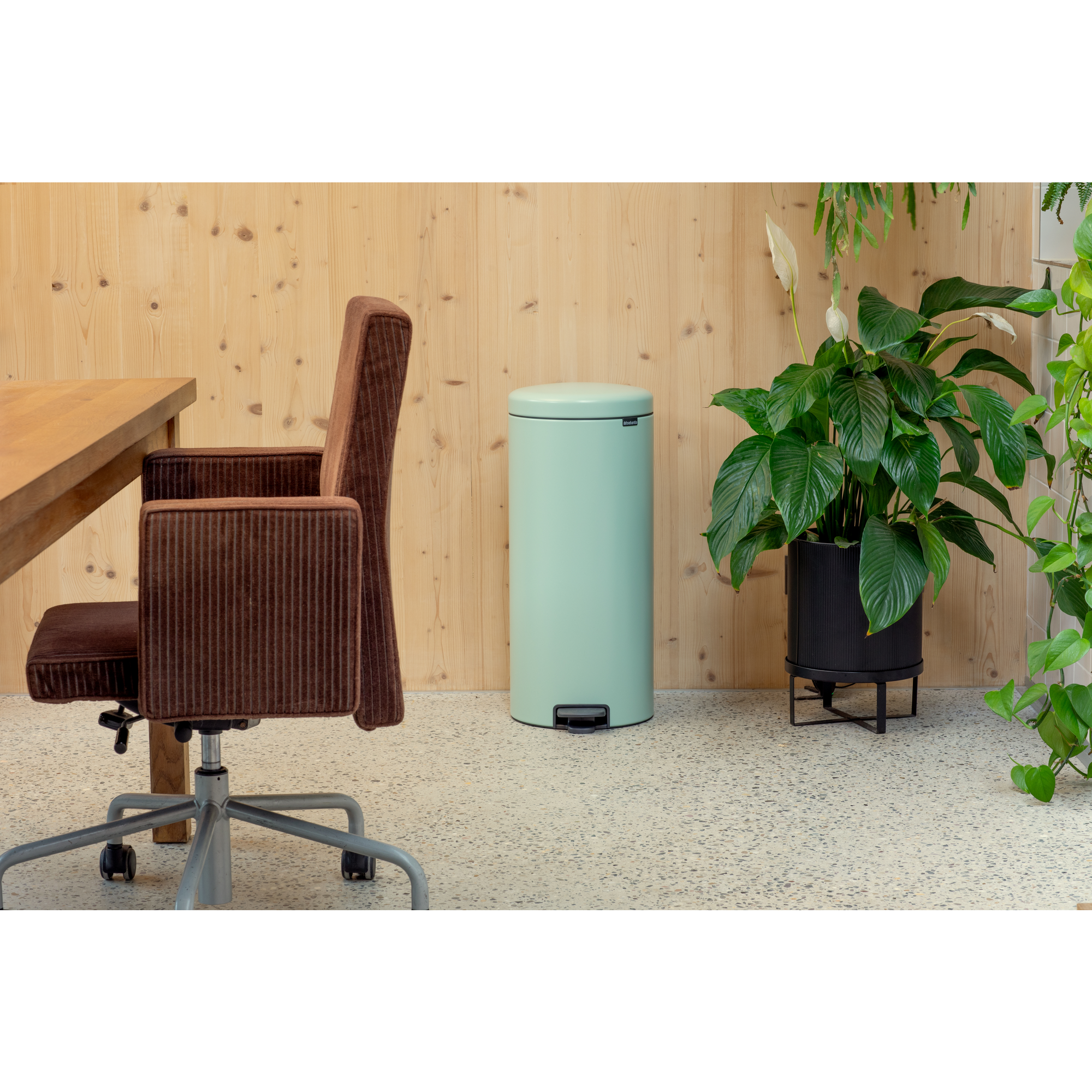 Una silla de oficina de pana marrón junto a una mesa de madera, un cubo de basura verde menta y plantas.