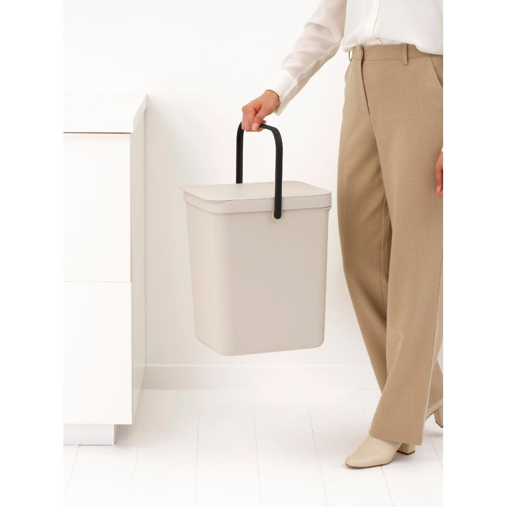 Een persoon in een beige broek en wit shirt houdt een beige prullenbak met zwarte handvatten vast.