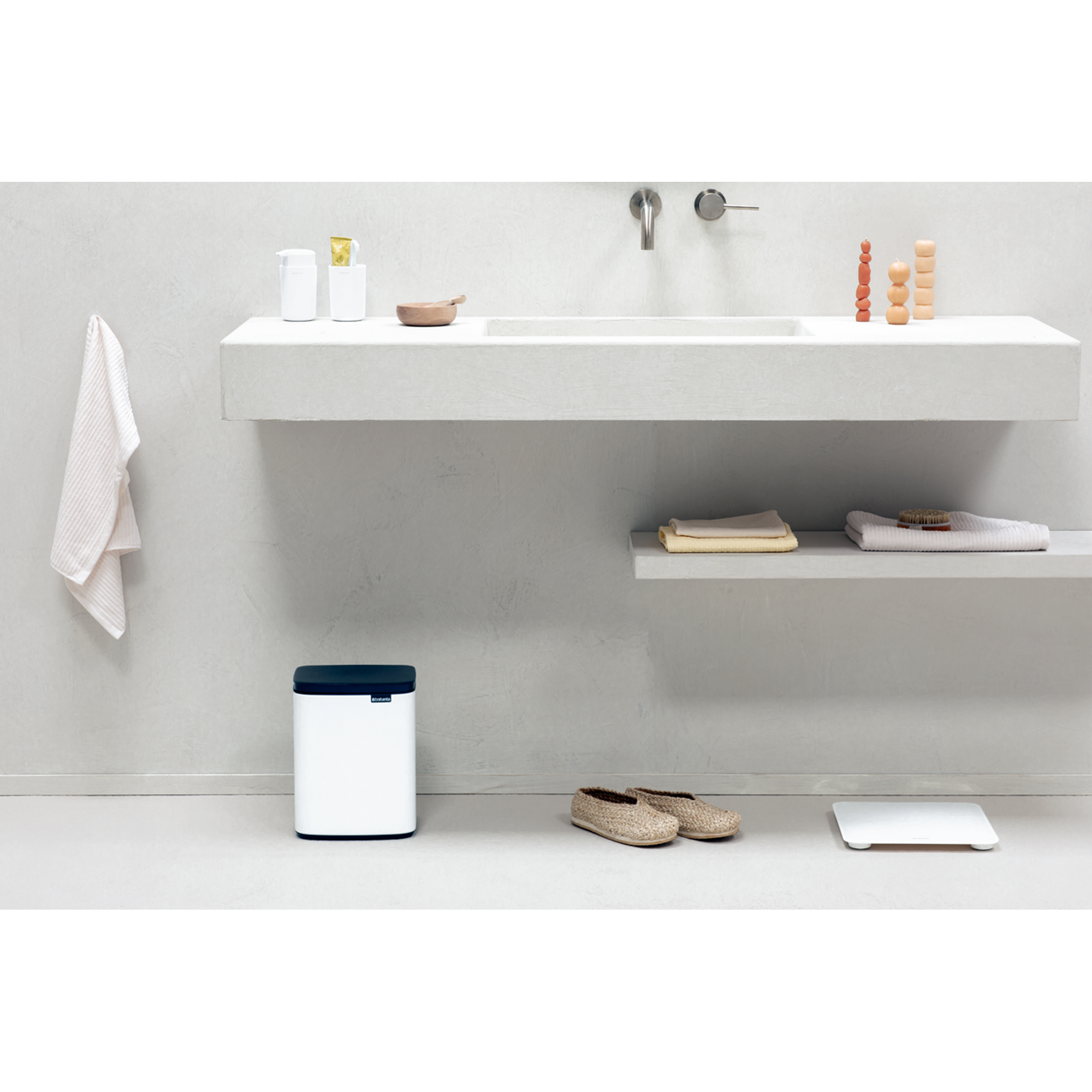 Minimalistische badkamer met witte prullenbak, weegschaal, slippers, handdoek en wastafel.
