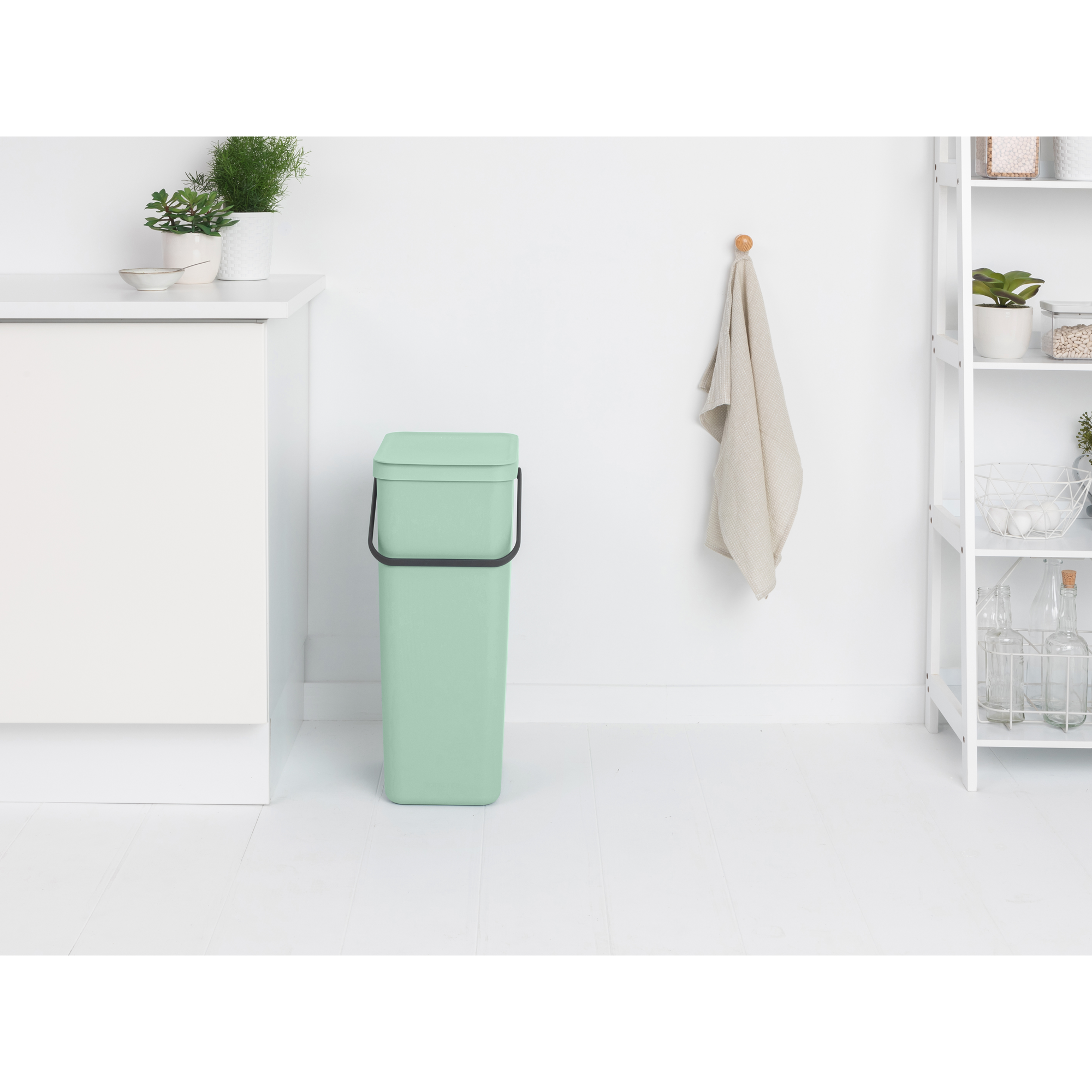 Une poubelle vert clair à côté d'une armoire blanche. Une serviette accrochée au mur.