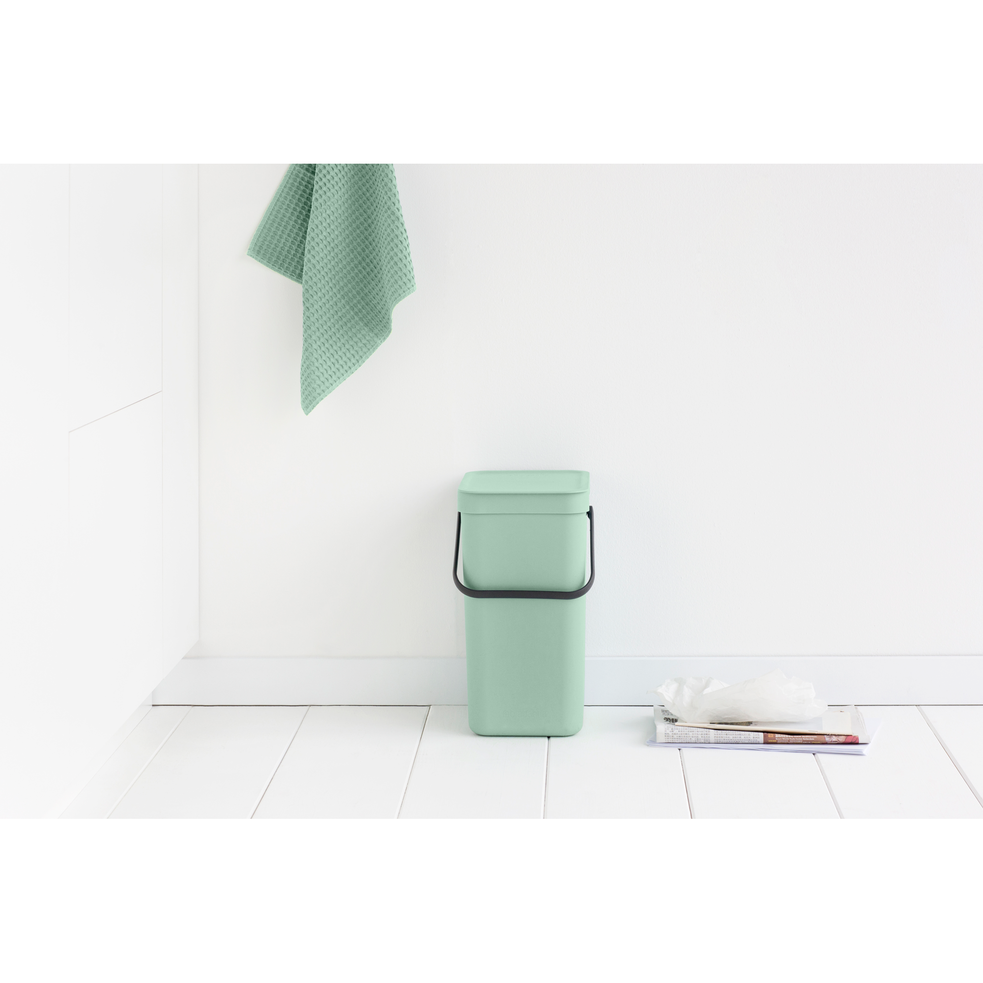 Een mintgroene bak met handvat staat op witte vloer, naast kranten.