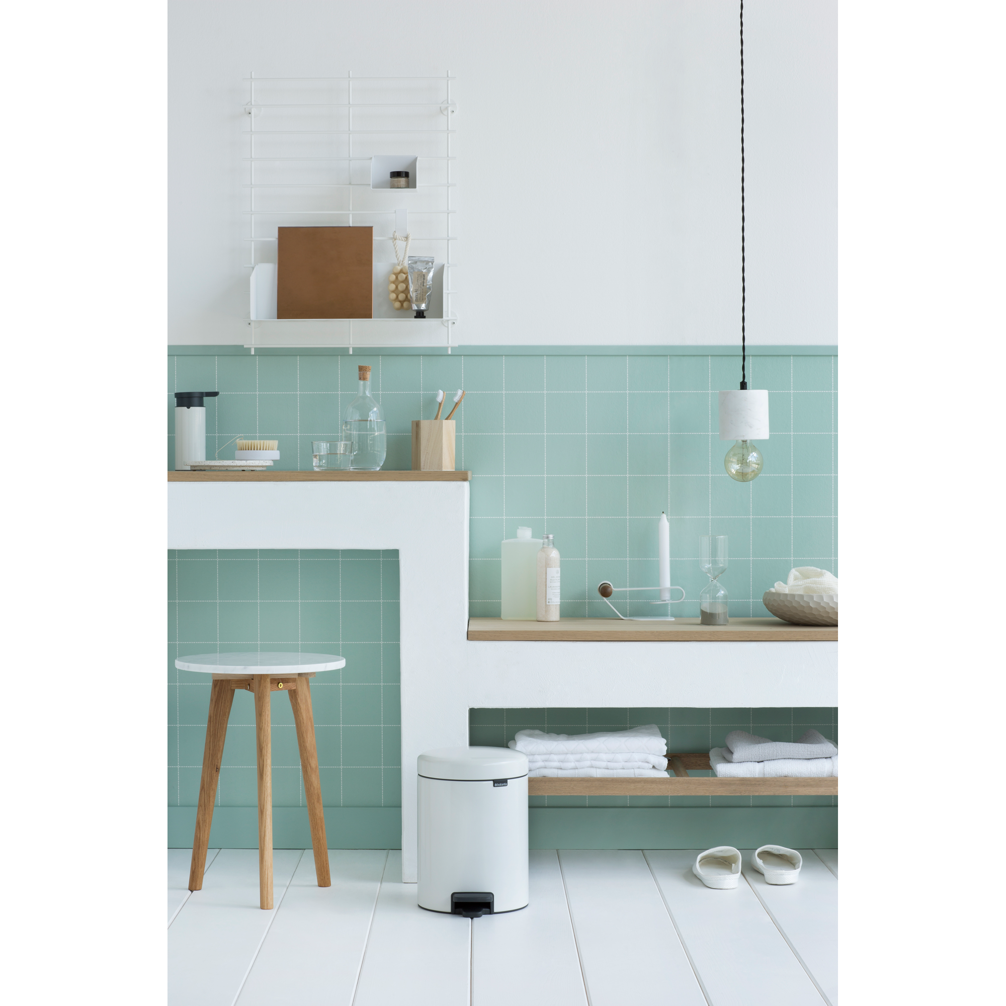 Een badkamer met groene tegels, een witte prullenbak, krukje en planken.