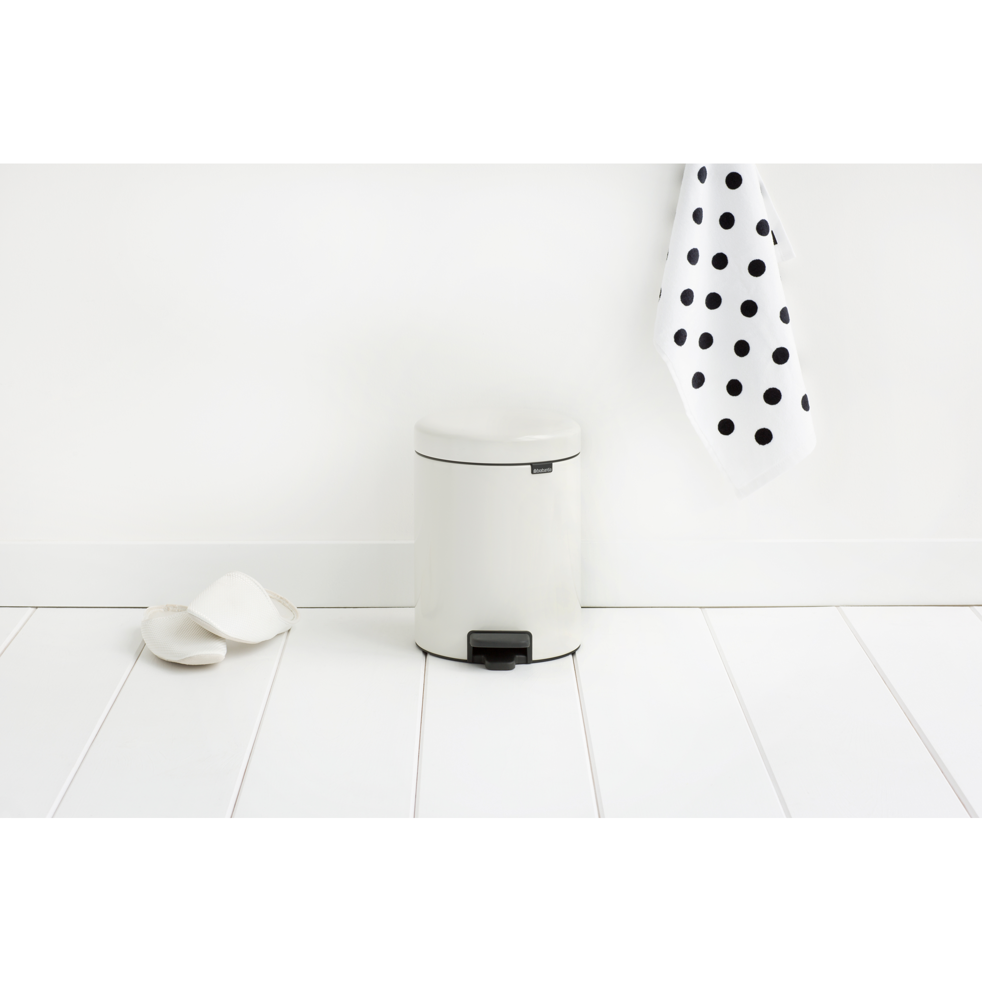 Een crème-kleurige prullenbak met pedaal staat op een witte houten vloer naast twee witte washandjes.
