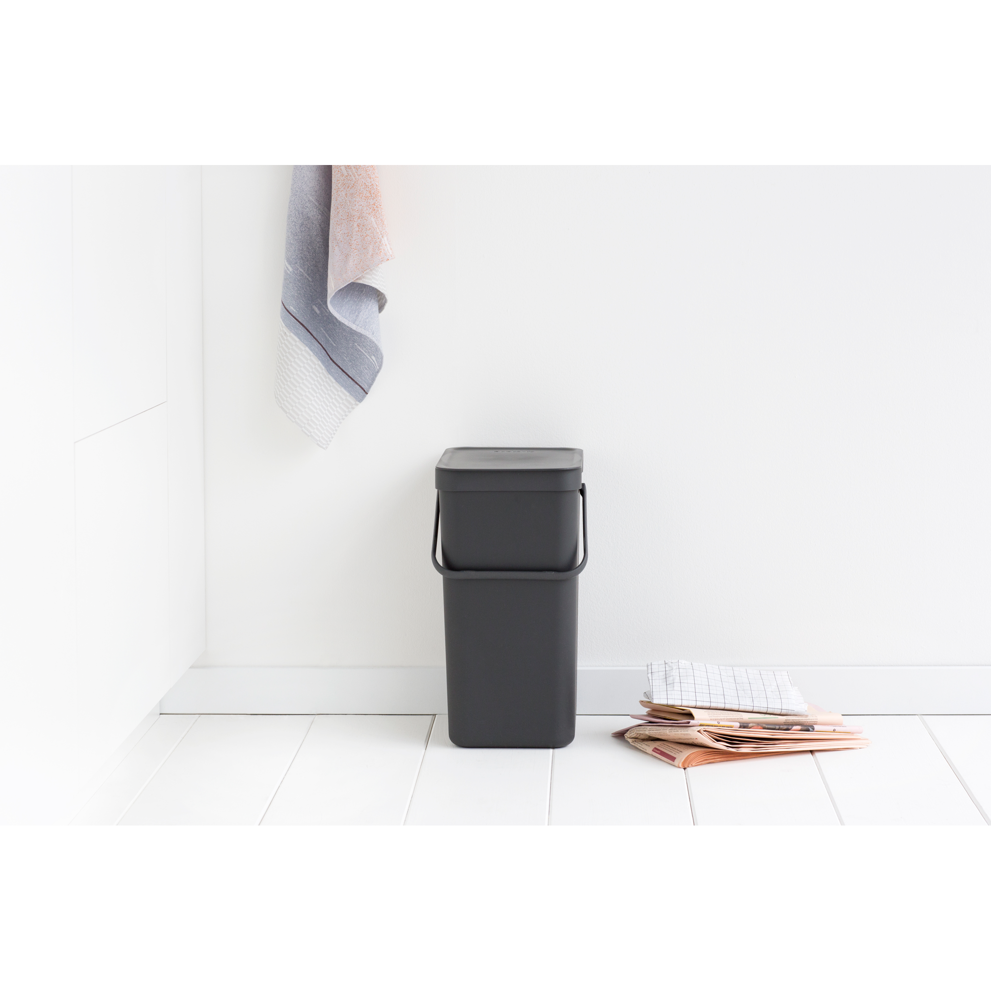 Een donkere vuilnisbak staat op een witte houten vloer naast kranten.