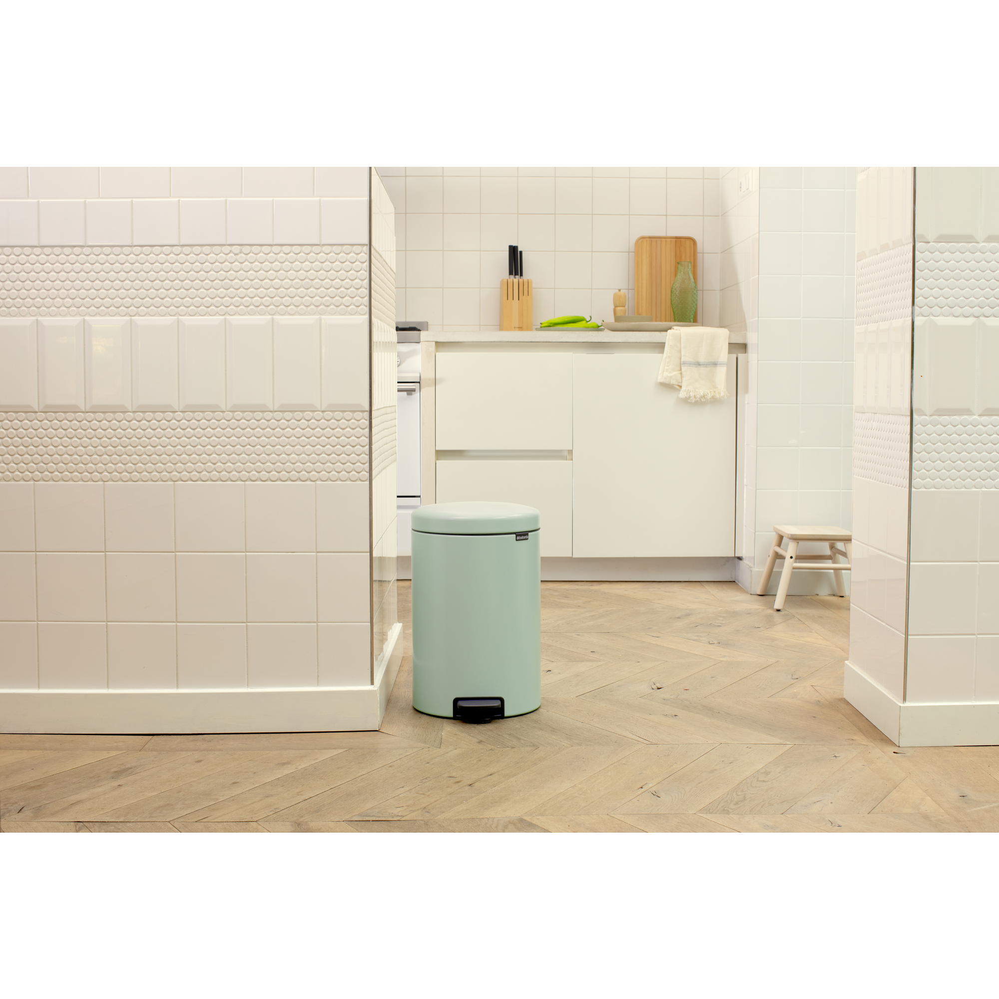 Un cubo de pedal verde menta sobre suelo de madera frente a una cocina blanca.