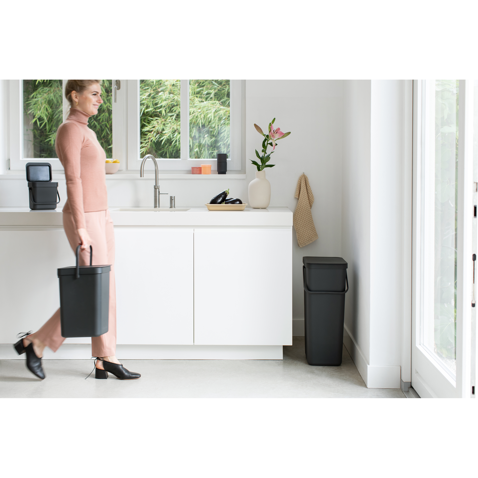 Een vrouw in het roze loopt met een grijze bak in een witte keuken met raam.