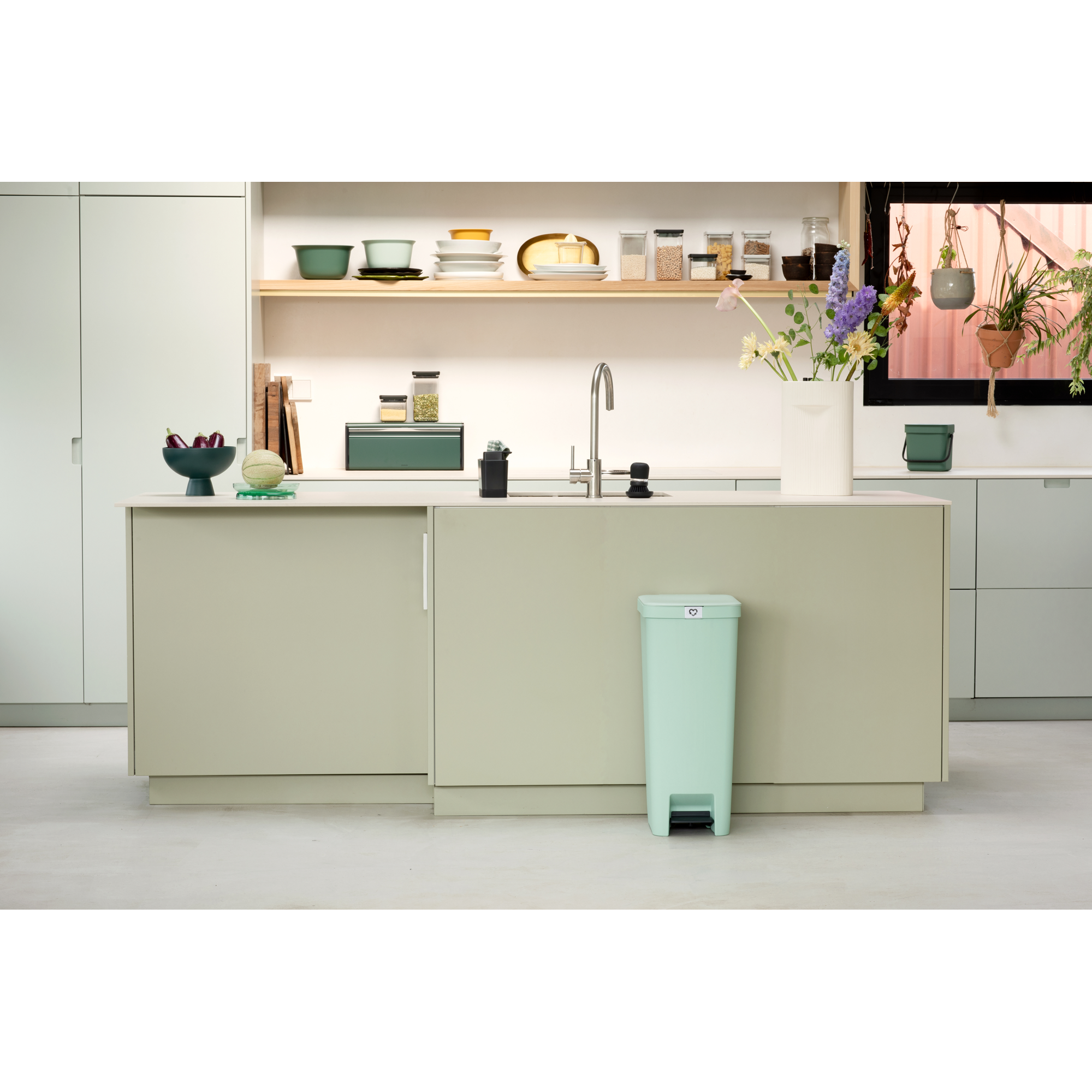 Een mintgroene prullenbak staat voor een lichtgroen keukeneiland met fruit en servies.
