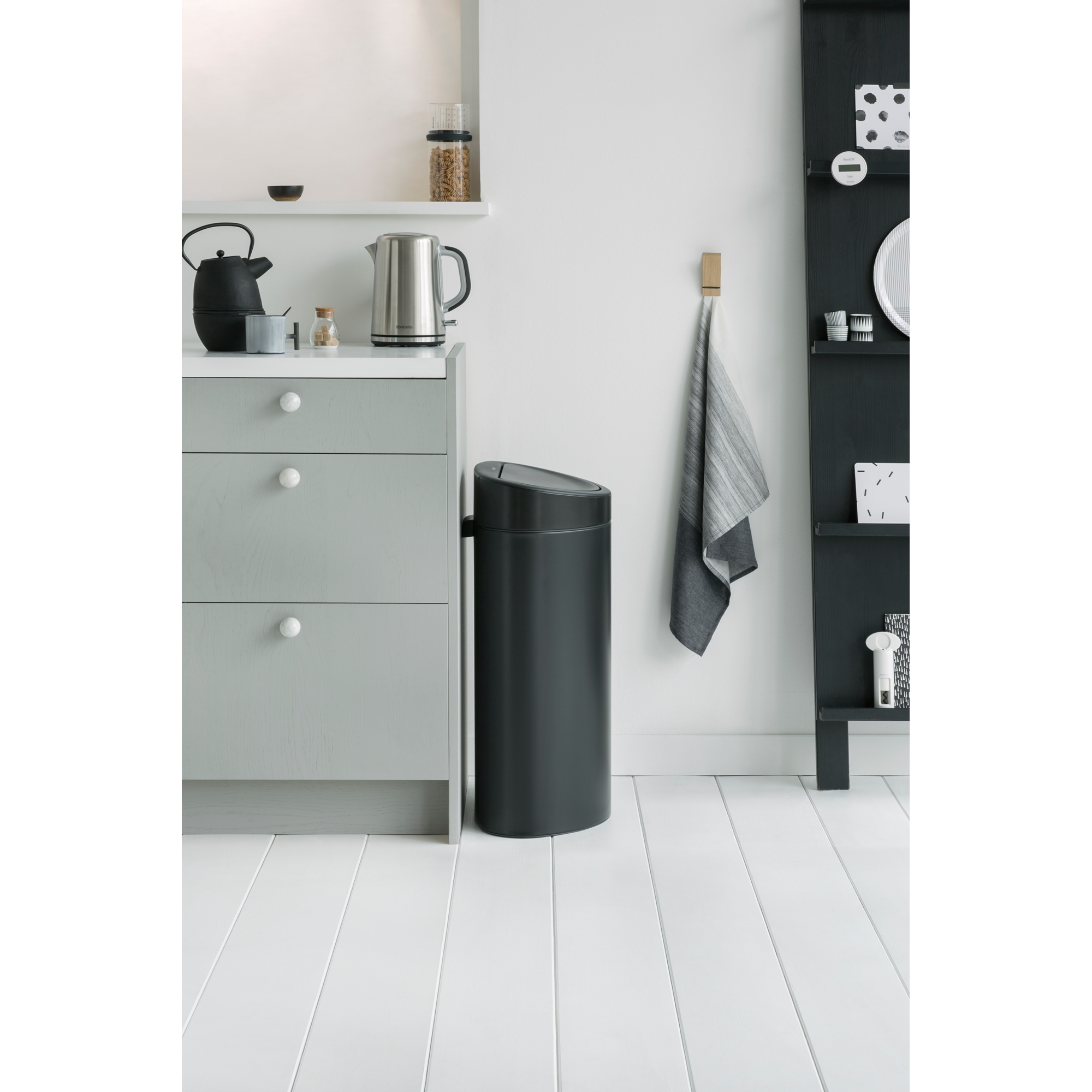 Een donkere prullenbak naast een lichtgrijze ladekast in een moderne keuken met witte vloerplanken.