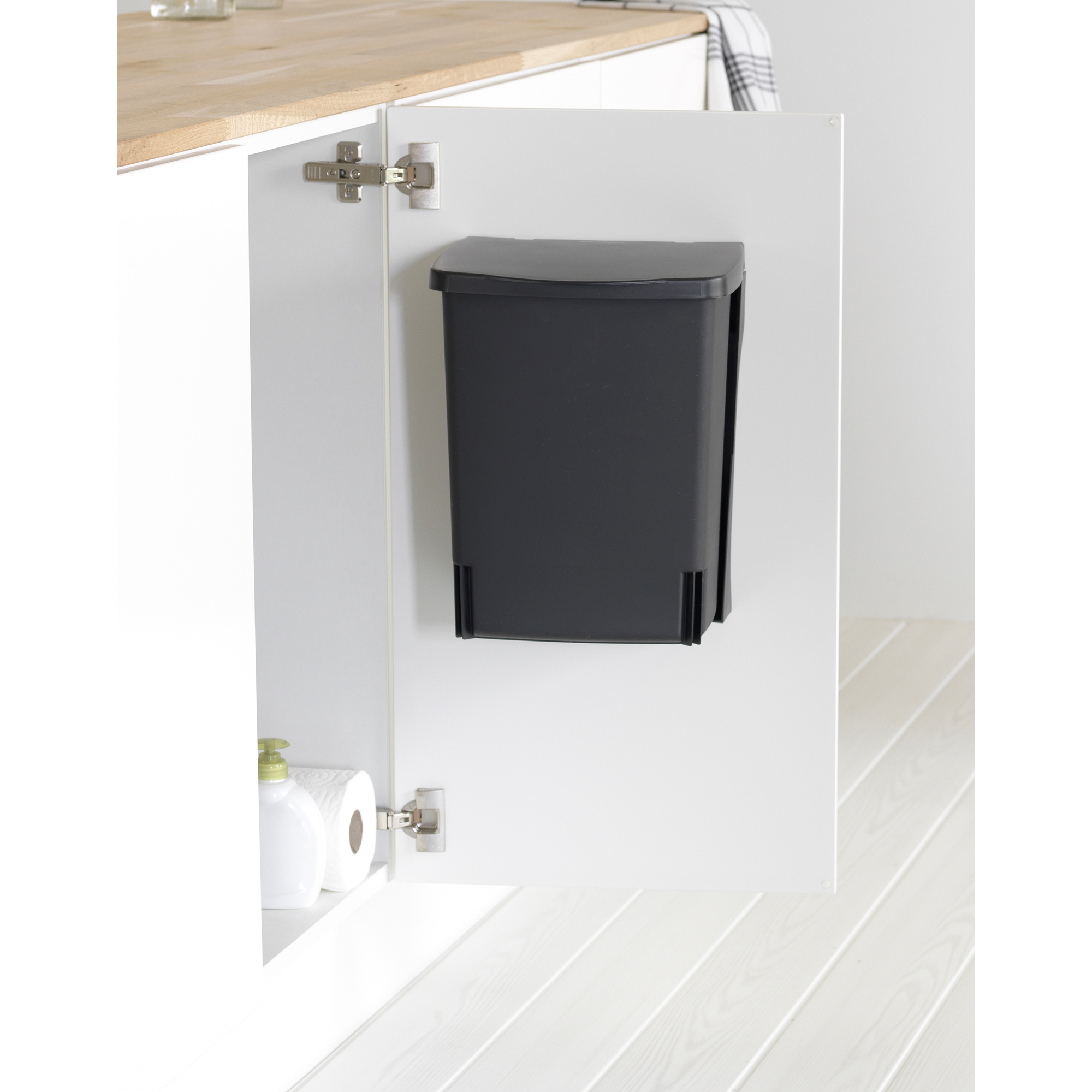 Donkergrijze prullenbak gemonteerd op witte kastdeur. Beneden zeep en toiletpapier.
