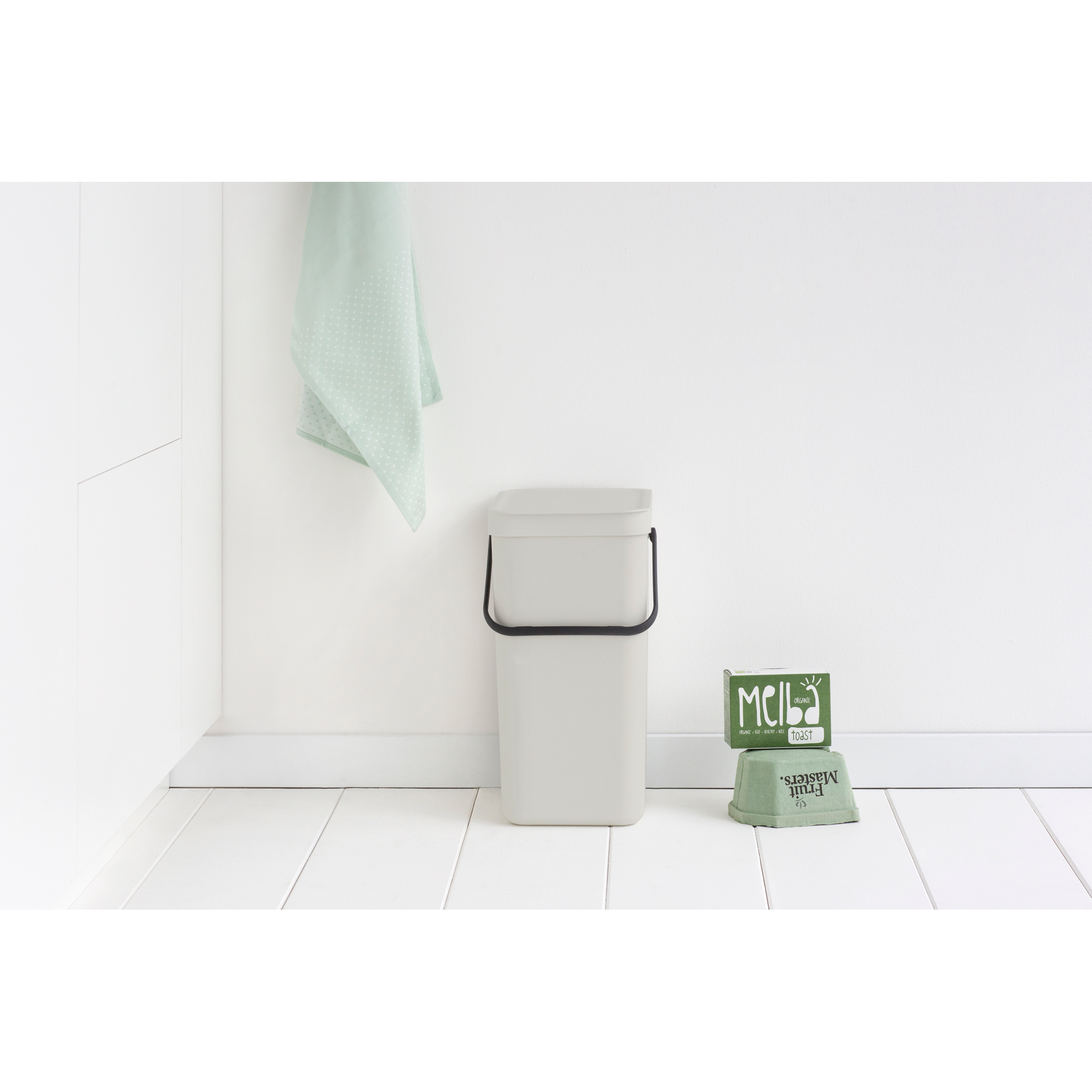 Een grijze prullenbak naast twee groene dozen op een witte houten vloer.