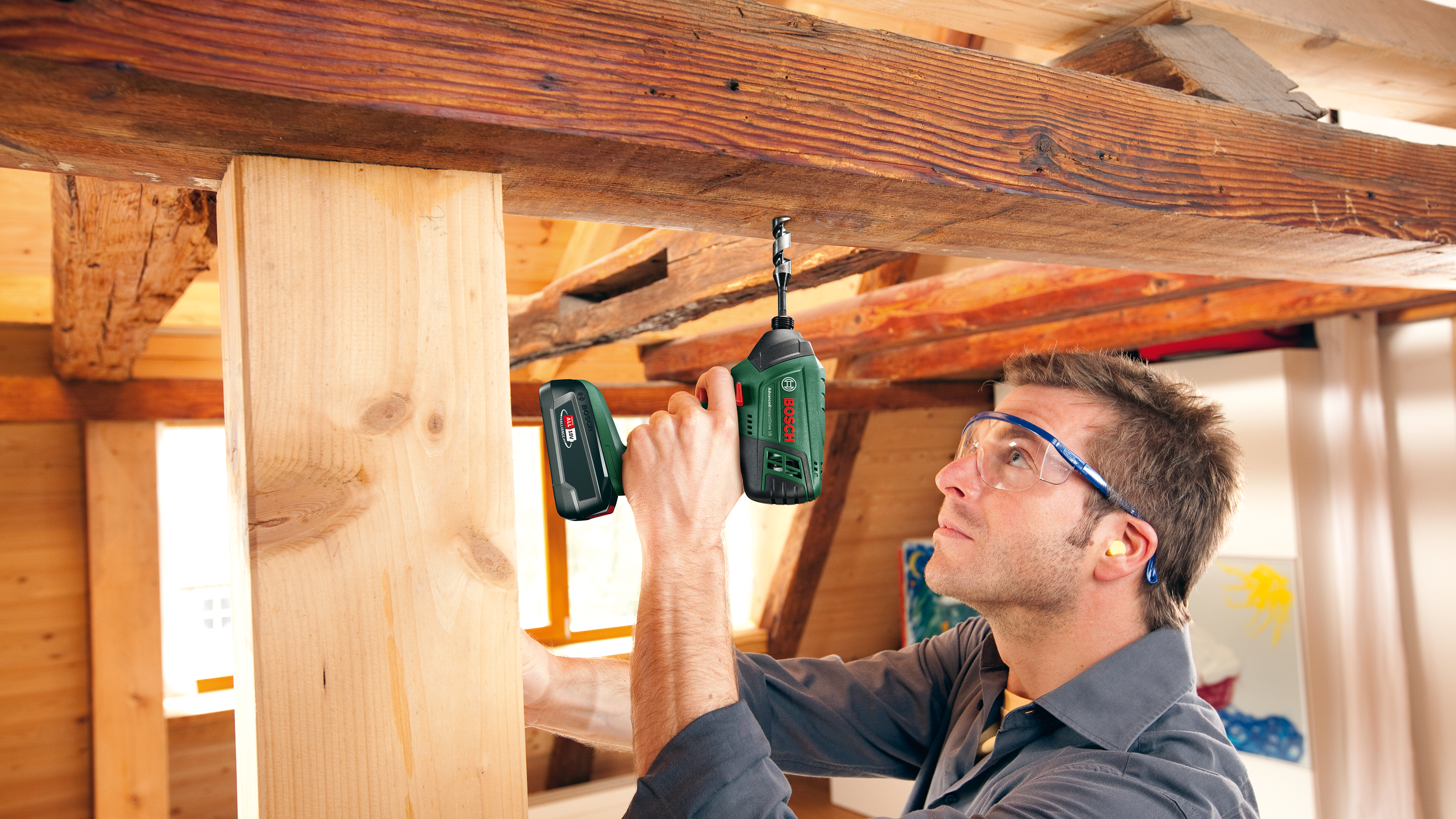 Mann mit Schutzbrille bohrt in Holzbalken in Werkstatt.
