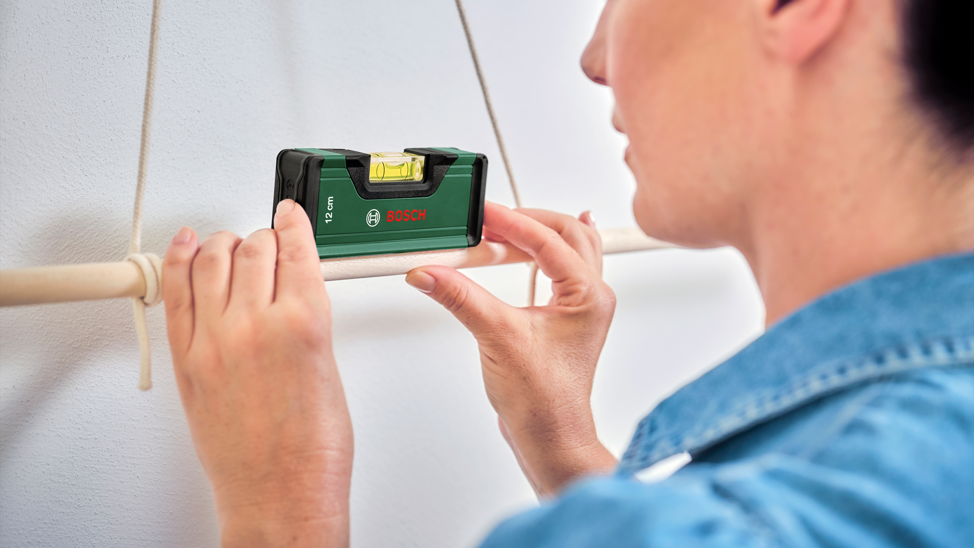 Bir kişi ipte asılı ahşap çubuğa Bosch su terazisi tutuyor.