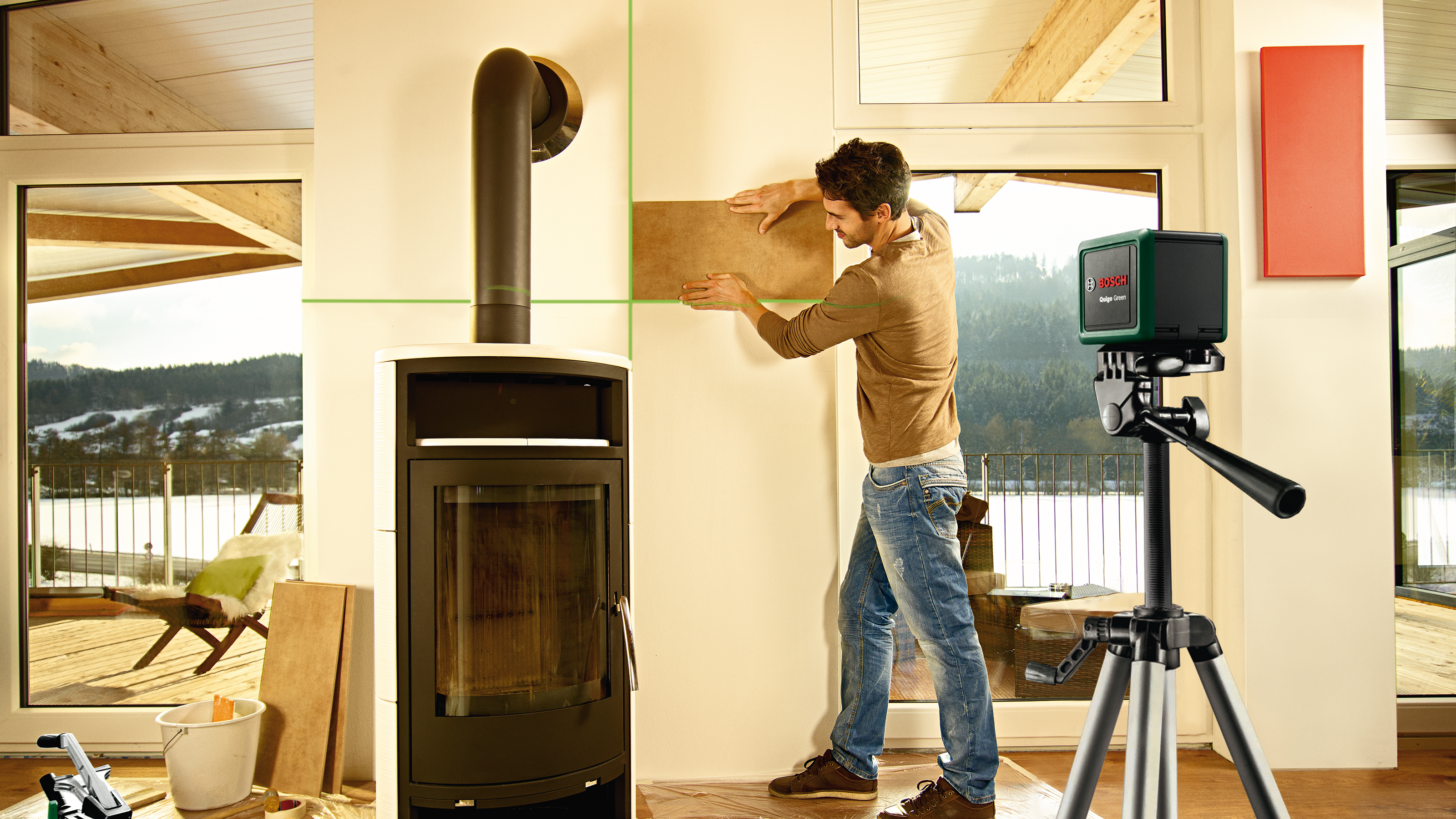 Man installs wall tiles with green laser level and tripod. Stove in room.