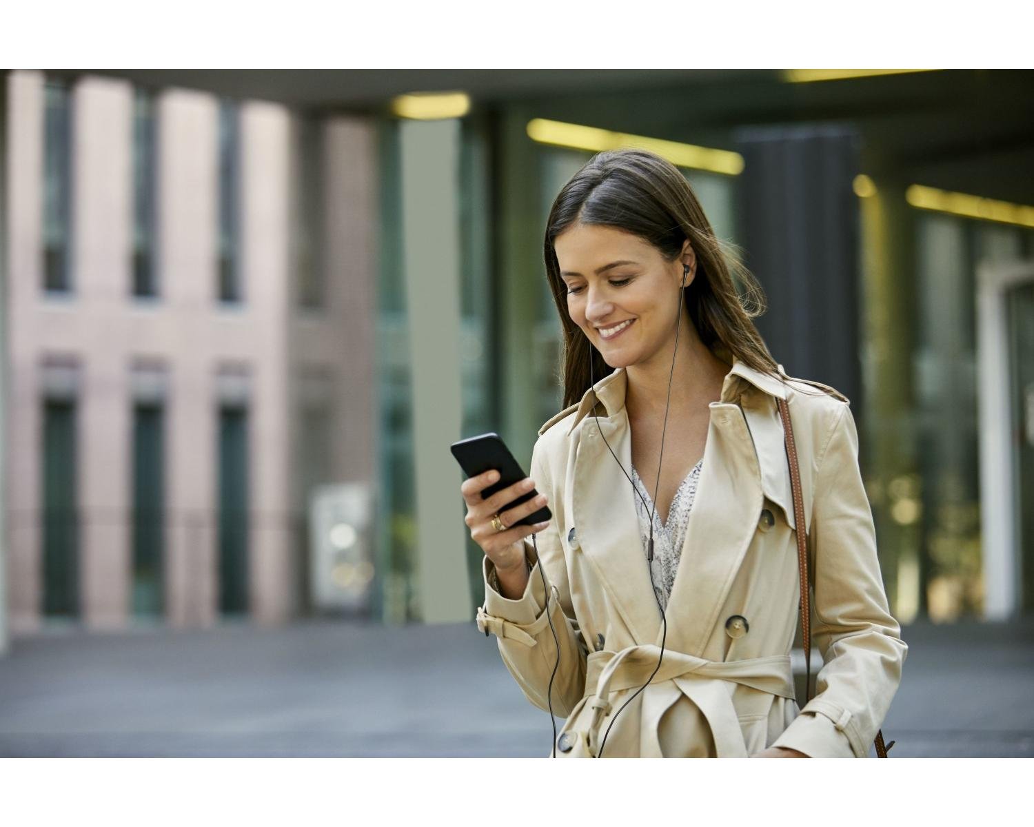 A woman in a beige trench coat smiles while looking at her smartphone.