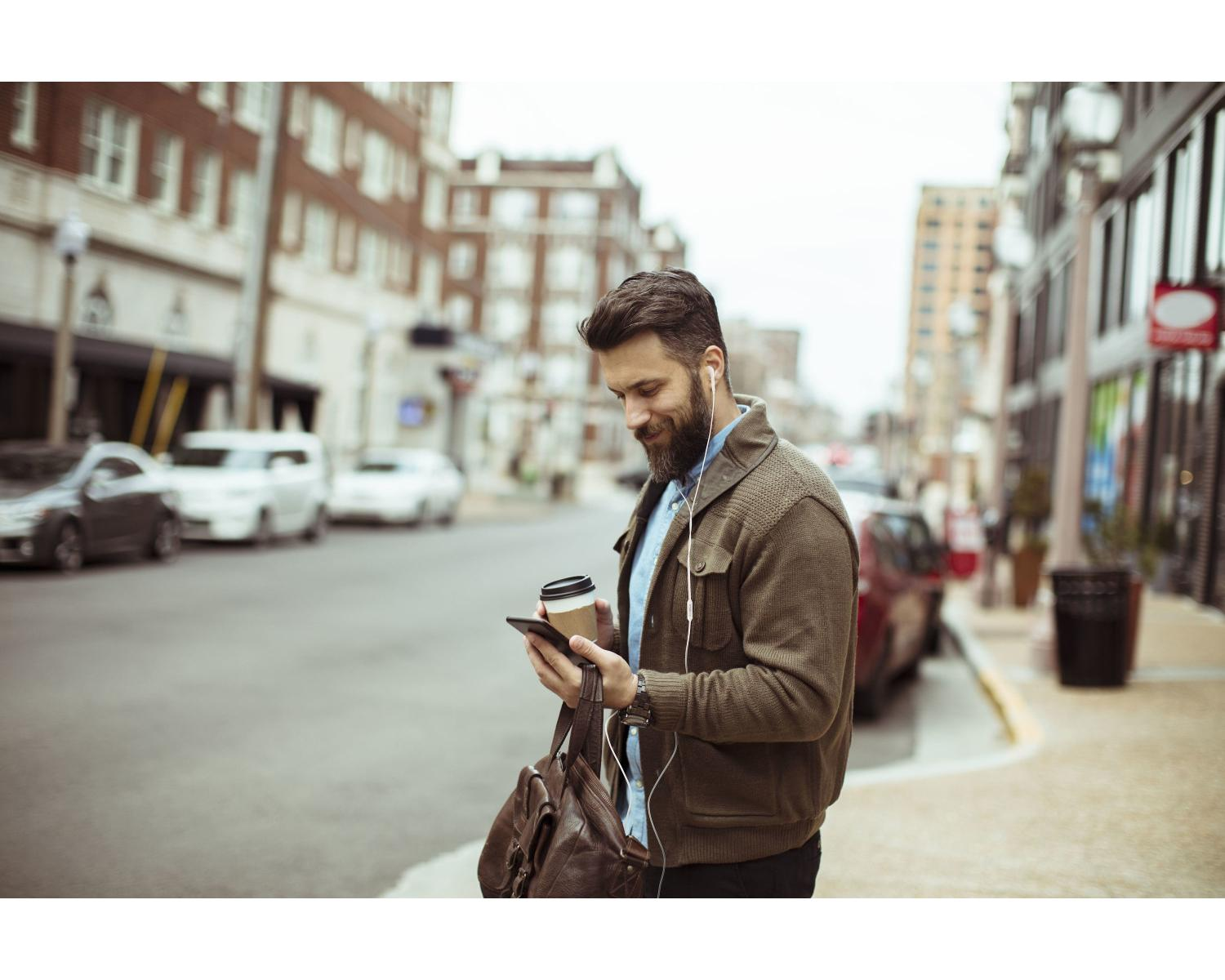 A man with a beard and headphones holds a phone and coffee cup on a city street.