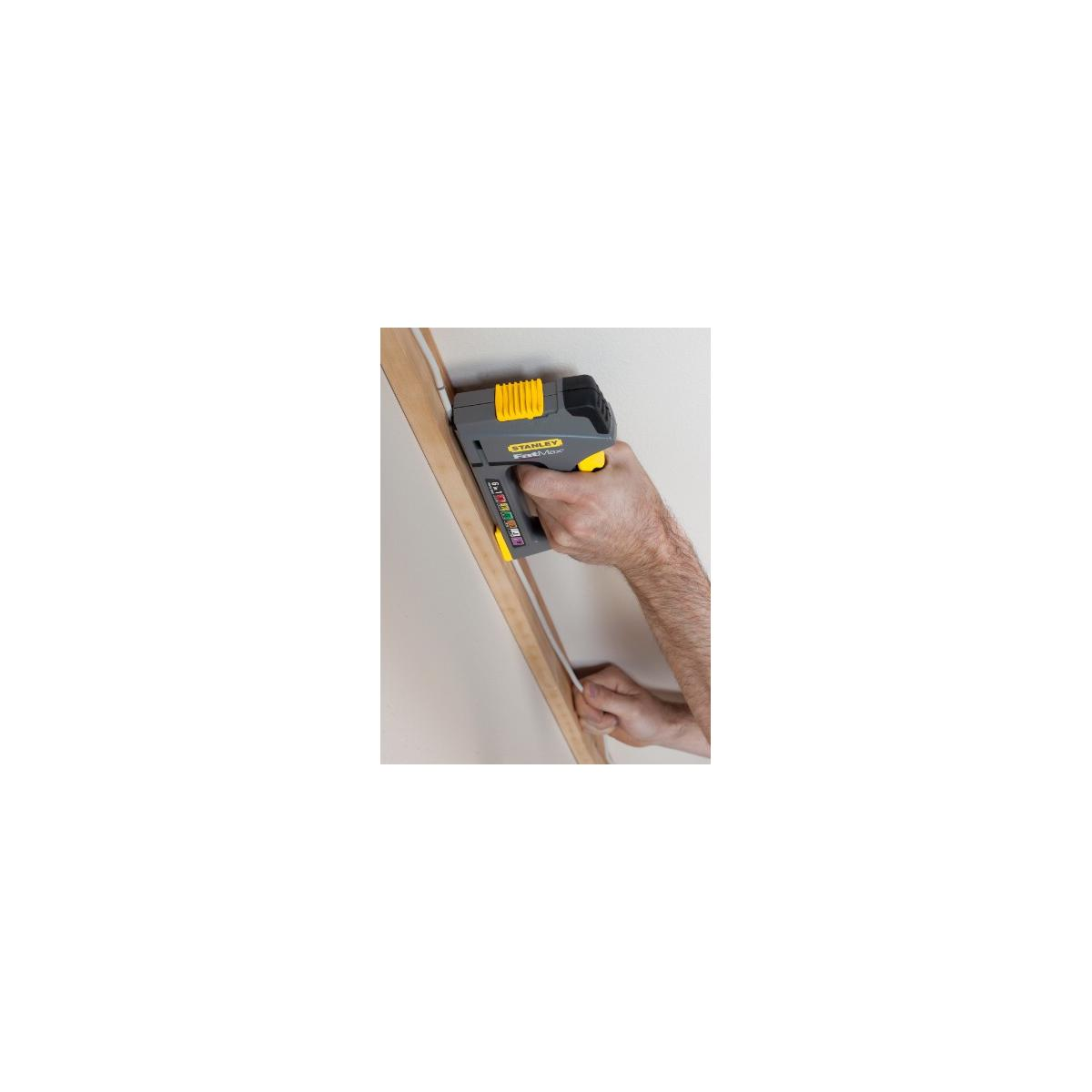 Grapadora gris y amarilla con mango amarillo, sujeta por mano, fijando tablón madera a pared.