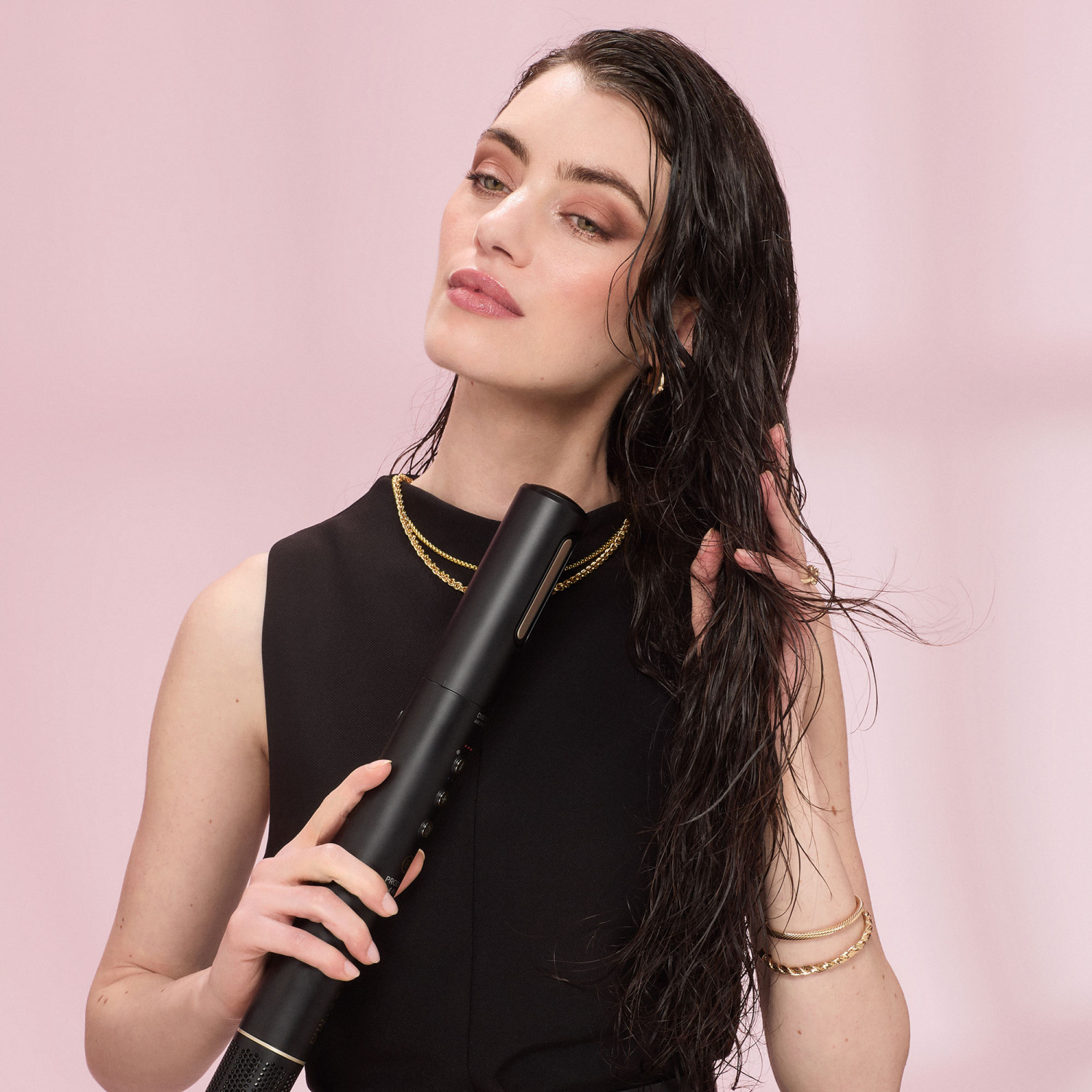 Donna con capelli bagnati tiene asciugacapelli nero, indossa abito nero e gioielli d'oro.