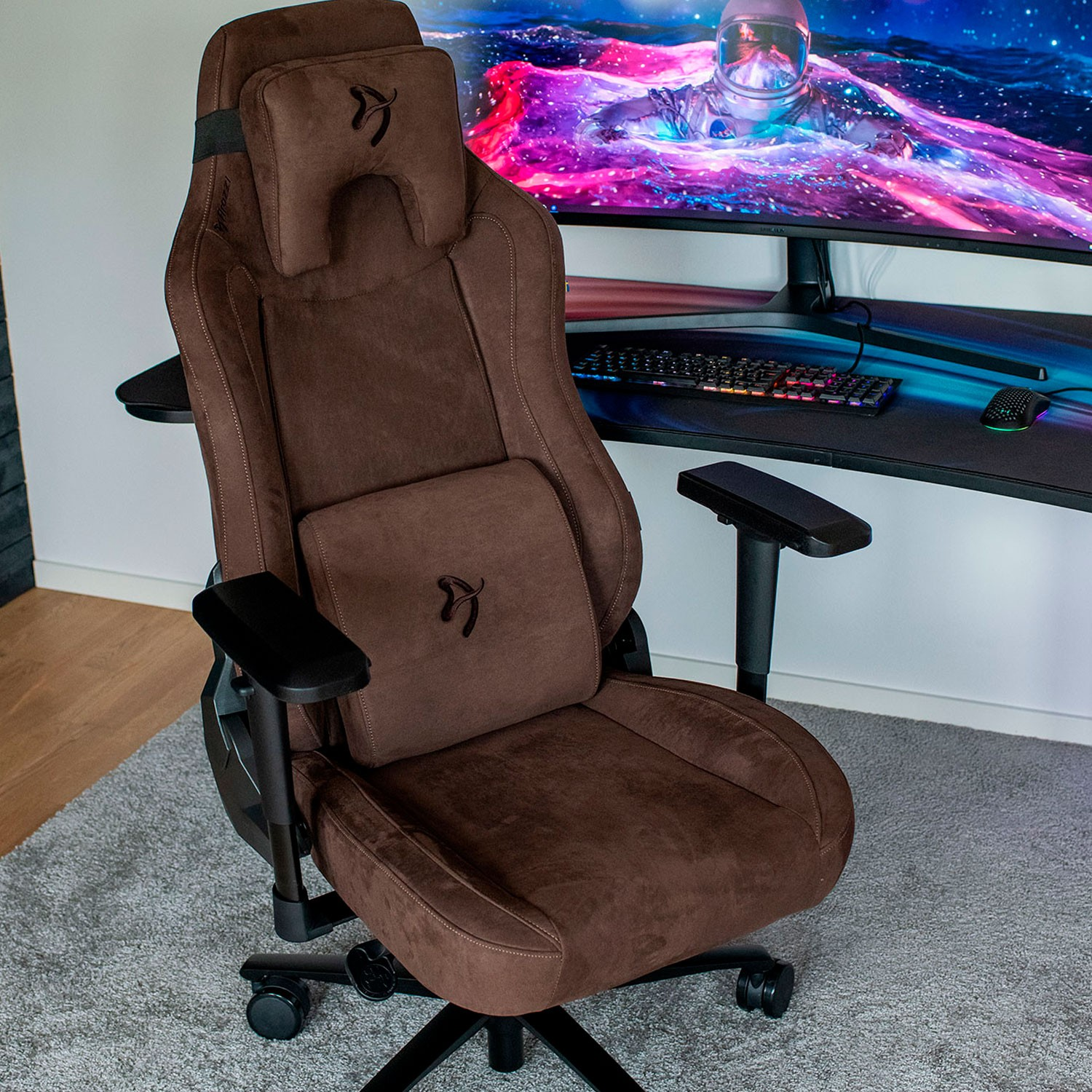 Ein brauner Gaming-Stuhl steht auf einem grauen Teppich vor einem Schreibtisch mit einem Monitor.