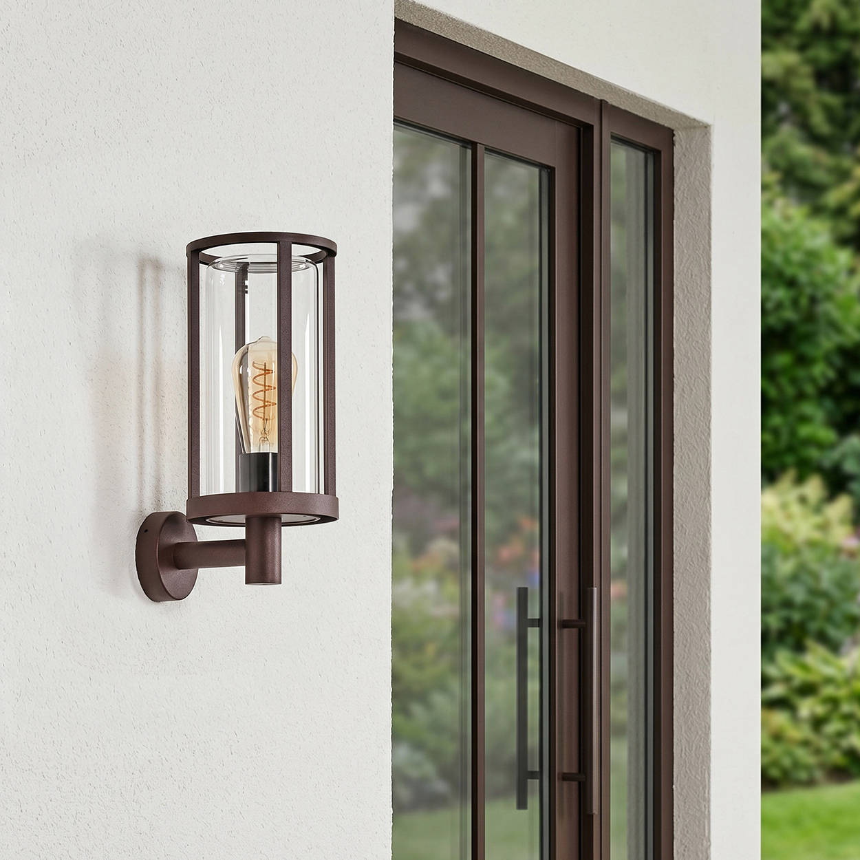 Eine braune Wandleuchte mit Glaszylinder und Glühbirne an einer weißen Wand neben einer Glastür.