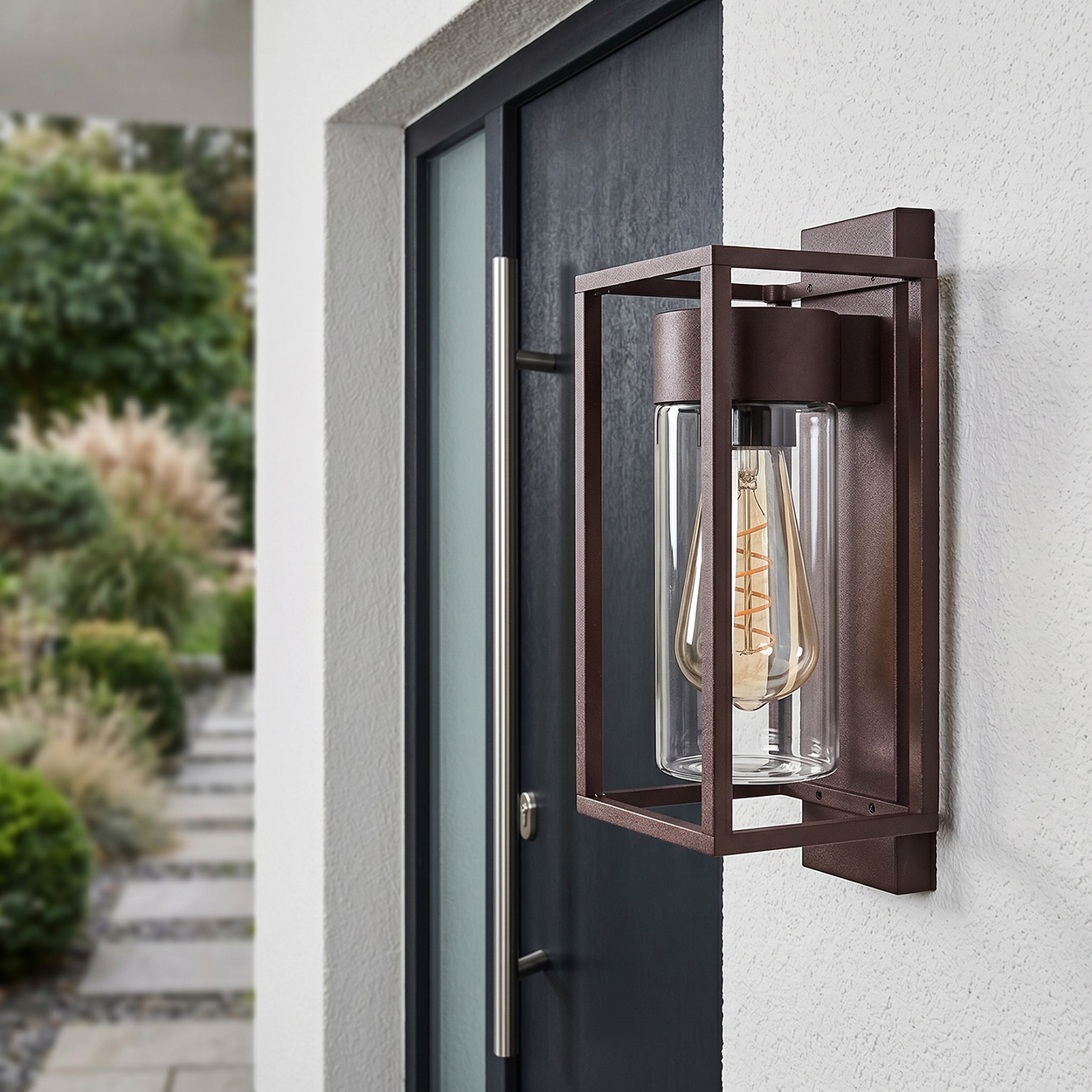 Eine braune Wandleuchte mit Glaszylinder und Glühbirne an einer weißen Wand neben einer Tür.