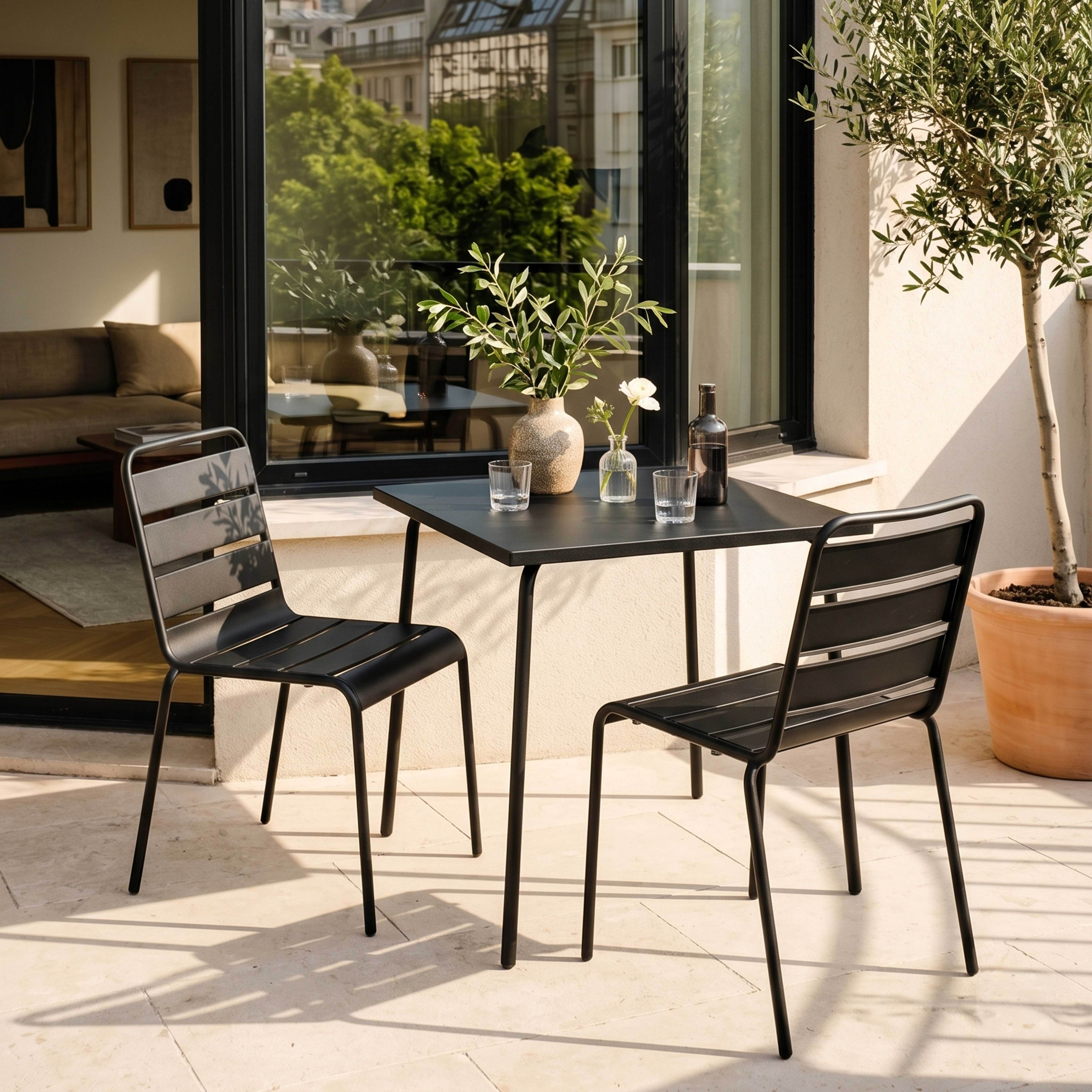 Schwarzer Tisch mit zwei Stühlen auf einer Terrasse mit Olivenbaum und Gebäuden im Hintergrund.