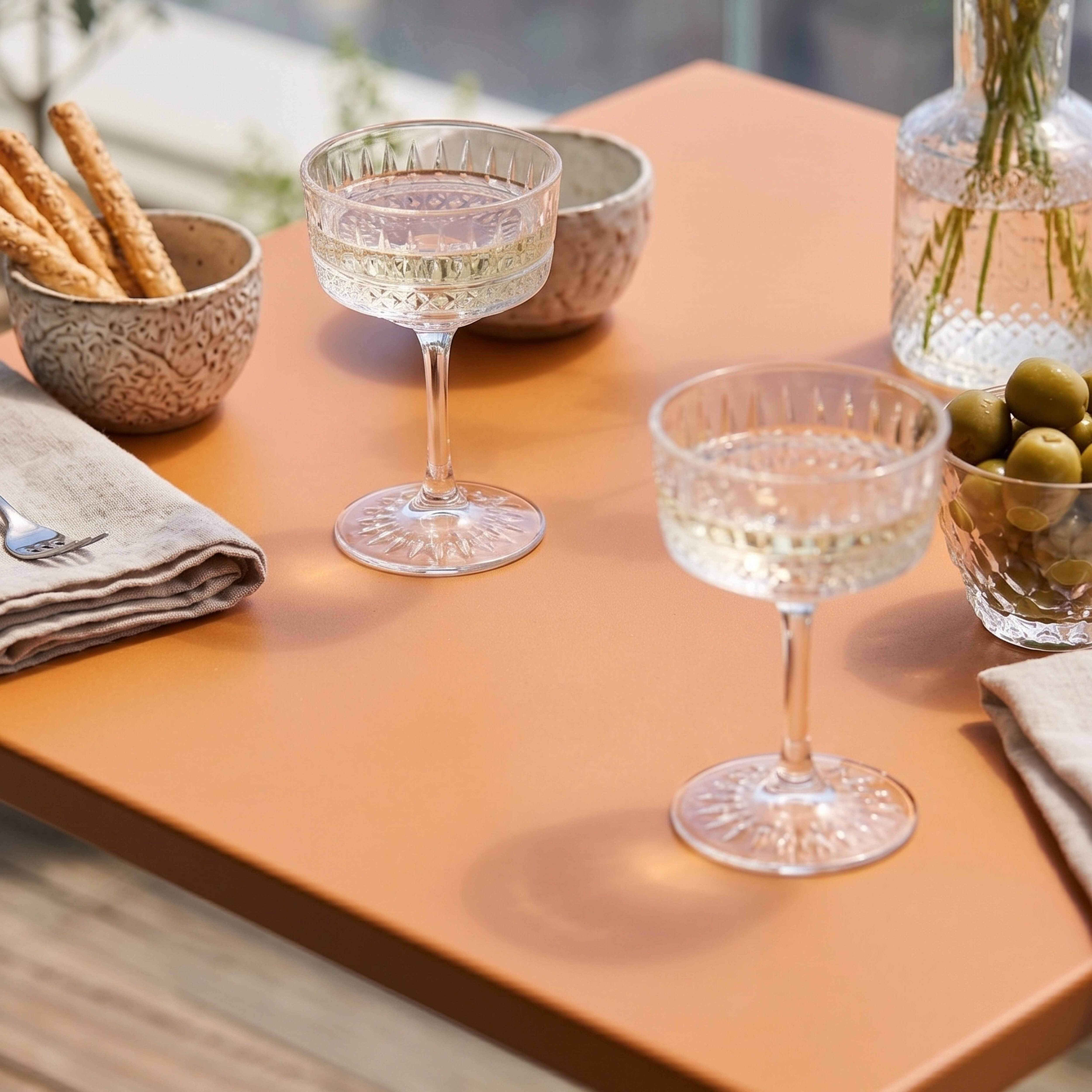 Zwei Gläser mit weißem Wein, Oliven, Grissini und einer Vase mit Pflanzen auf einem Tisch.