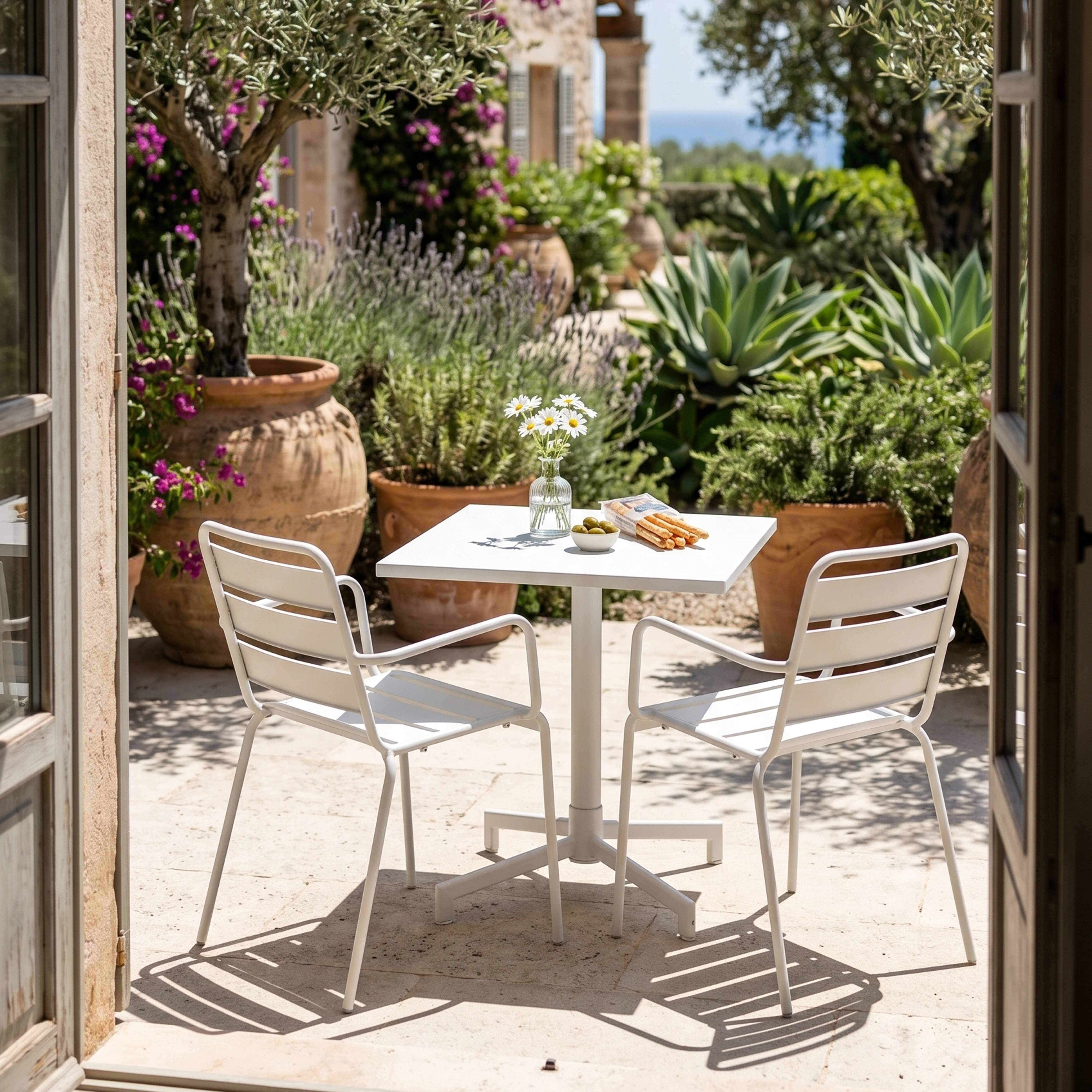 Ein weißer Tisch mit zwei Stühlen steht in einem sonnigen Garten mit Olivenbäumen.