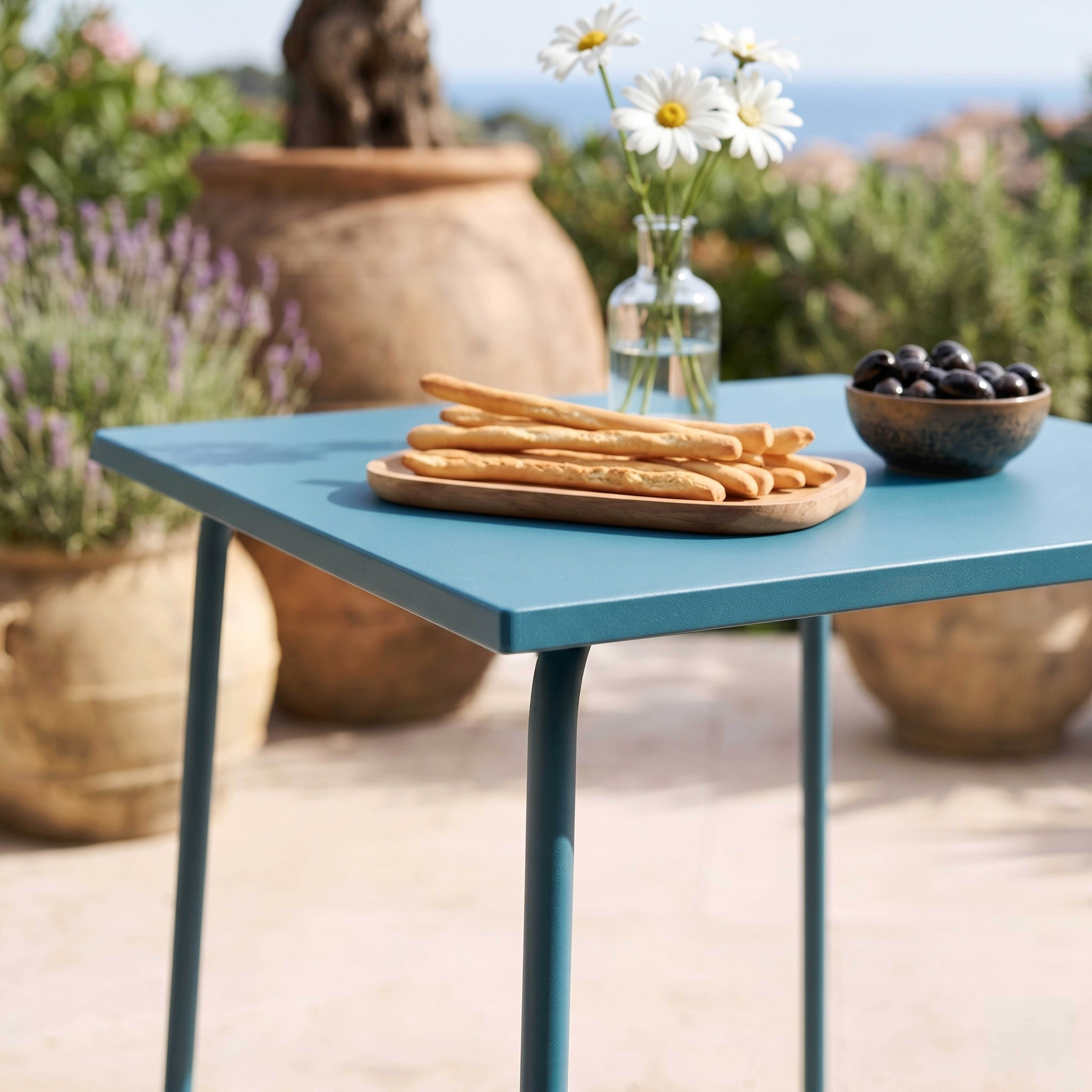 Ein blauer Tisch im Freien mit Brotstangen, Oliven, Gänseblümchen und Pflanzen.