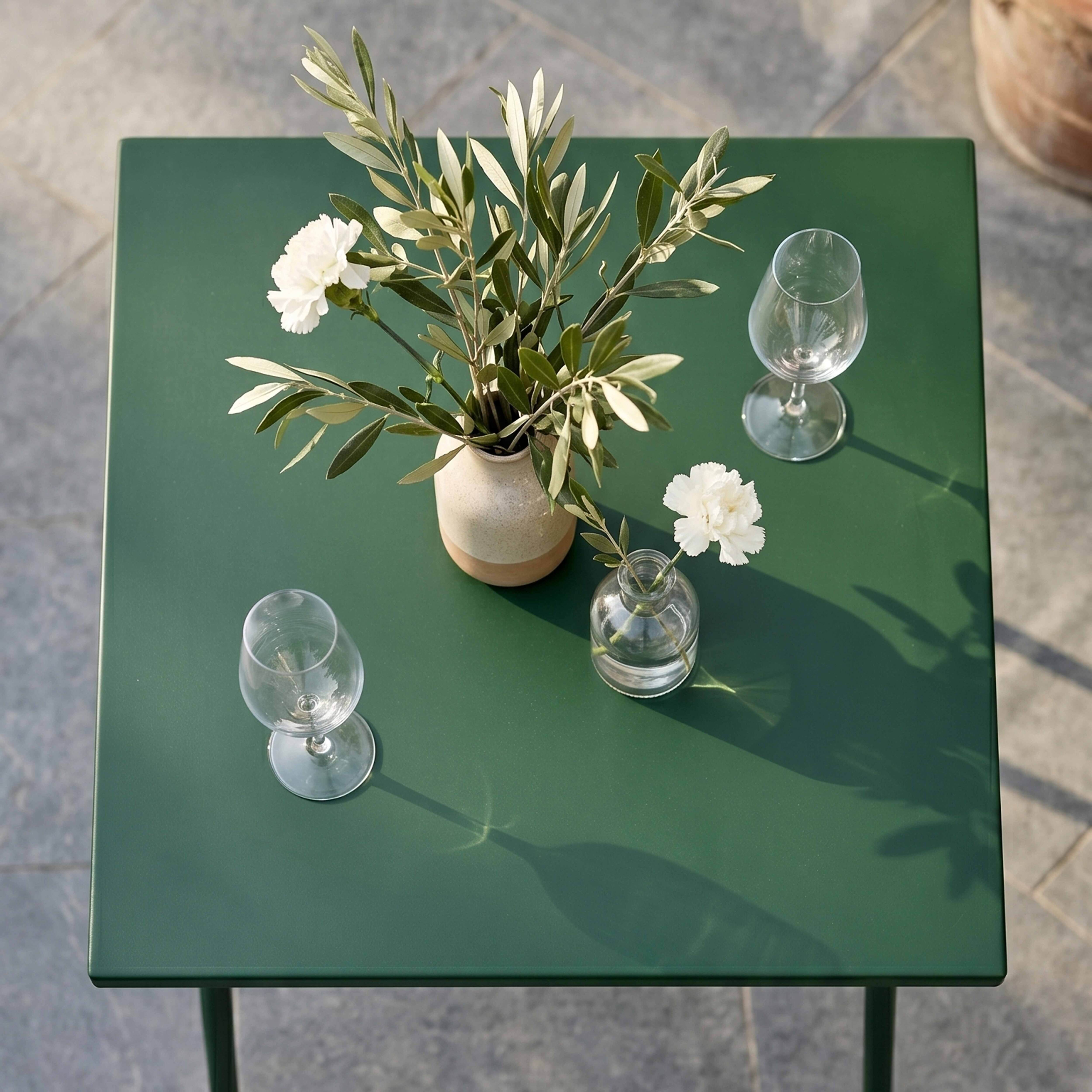 Ein grüner Tisch mit zwei Weingläsern, einer Vase mit Olivenzweigen und einer Blüte.