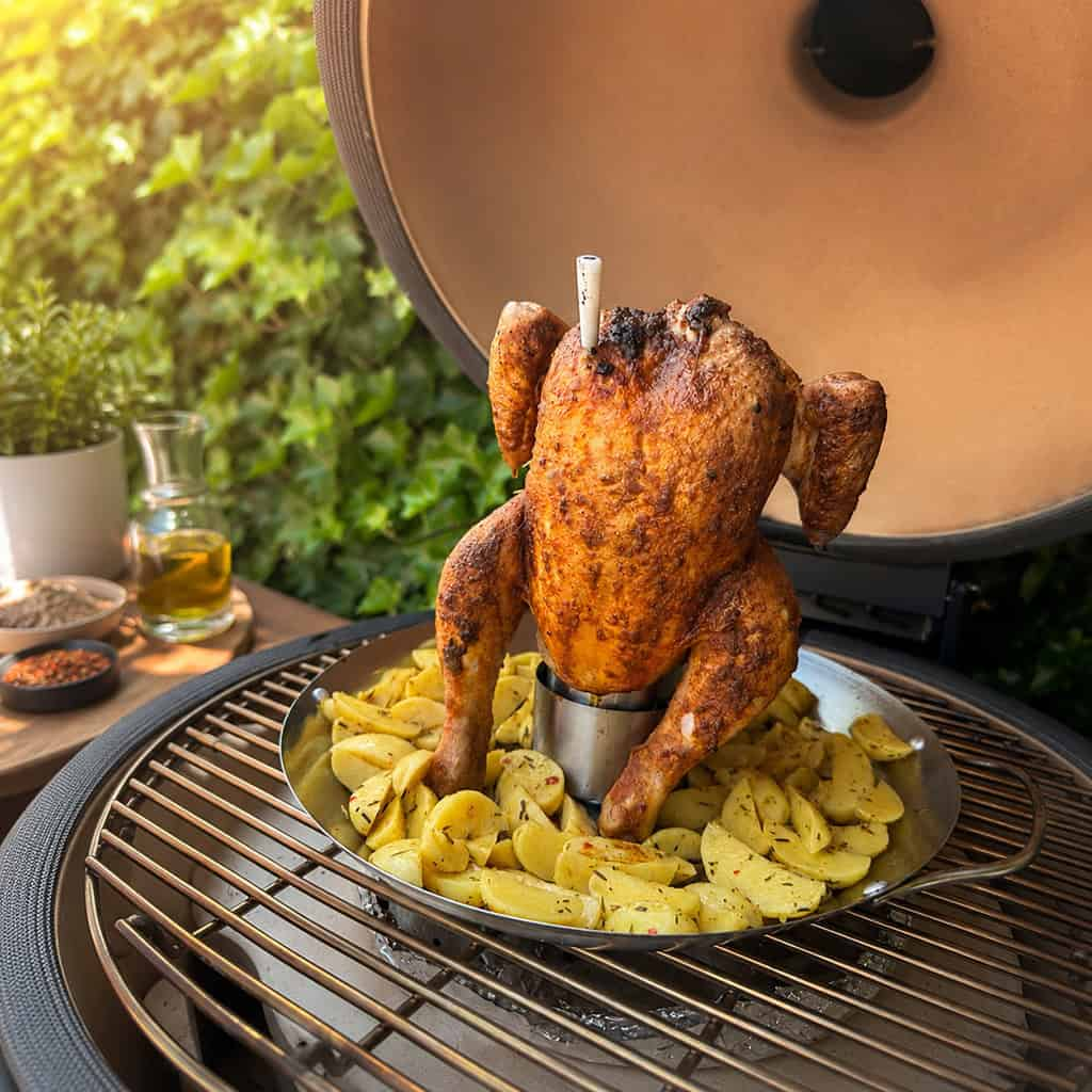 A golden-brown roasted chicken stands on potato wedges in a grill.
