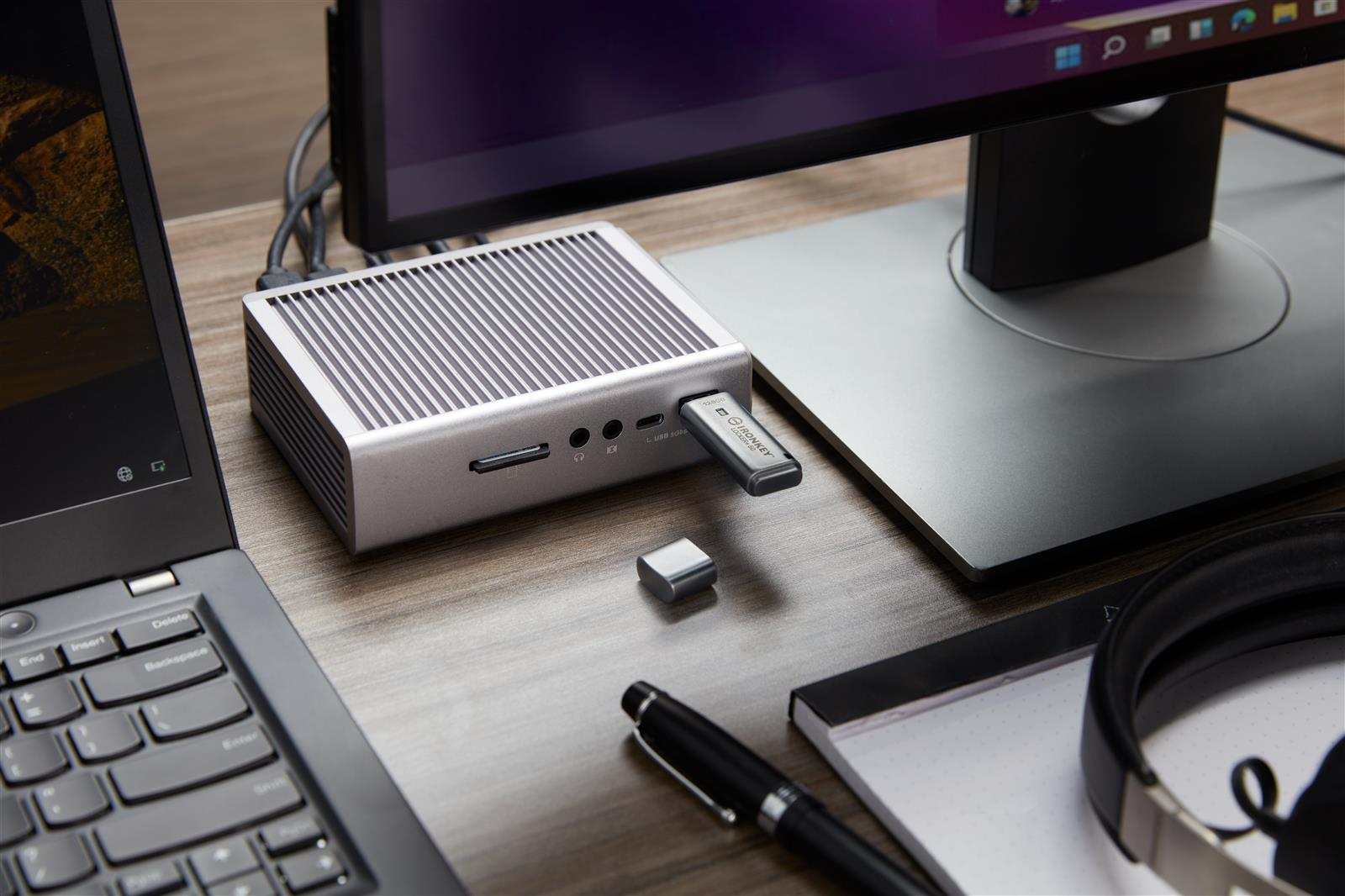 A silver mini PC with USB drive, laptop, monitor, headphones, pen and notepad.