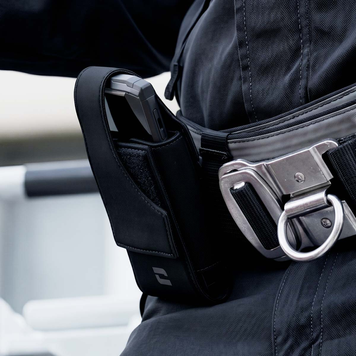 Primer plano de un cinturón utilitario negro con funda para teléfono y gran hebilla metálica.