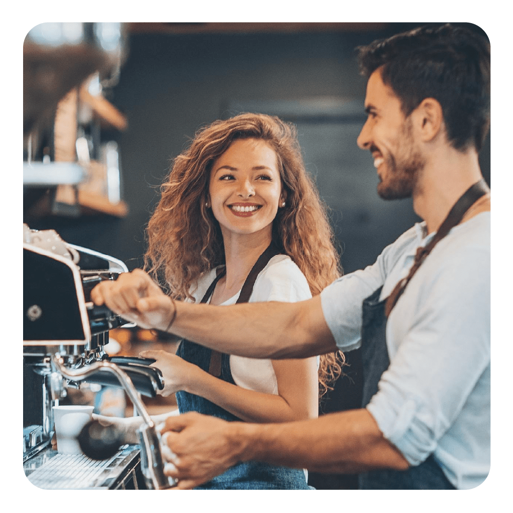 Un uomo e una donna con grembiuli si sorridono mentre preparano il caffè.