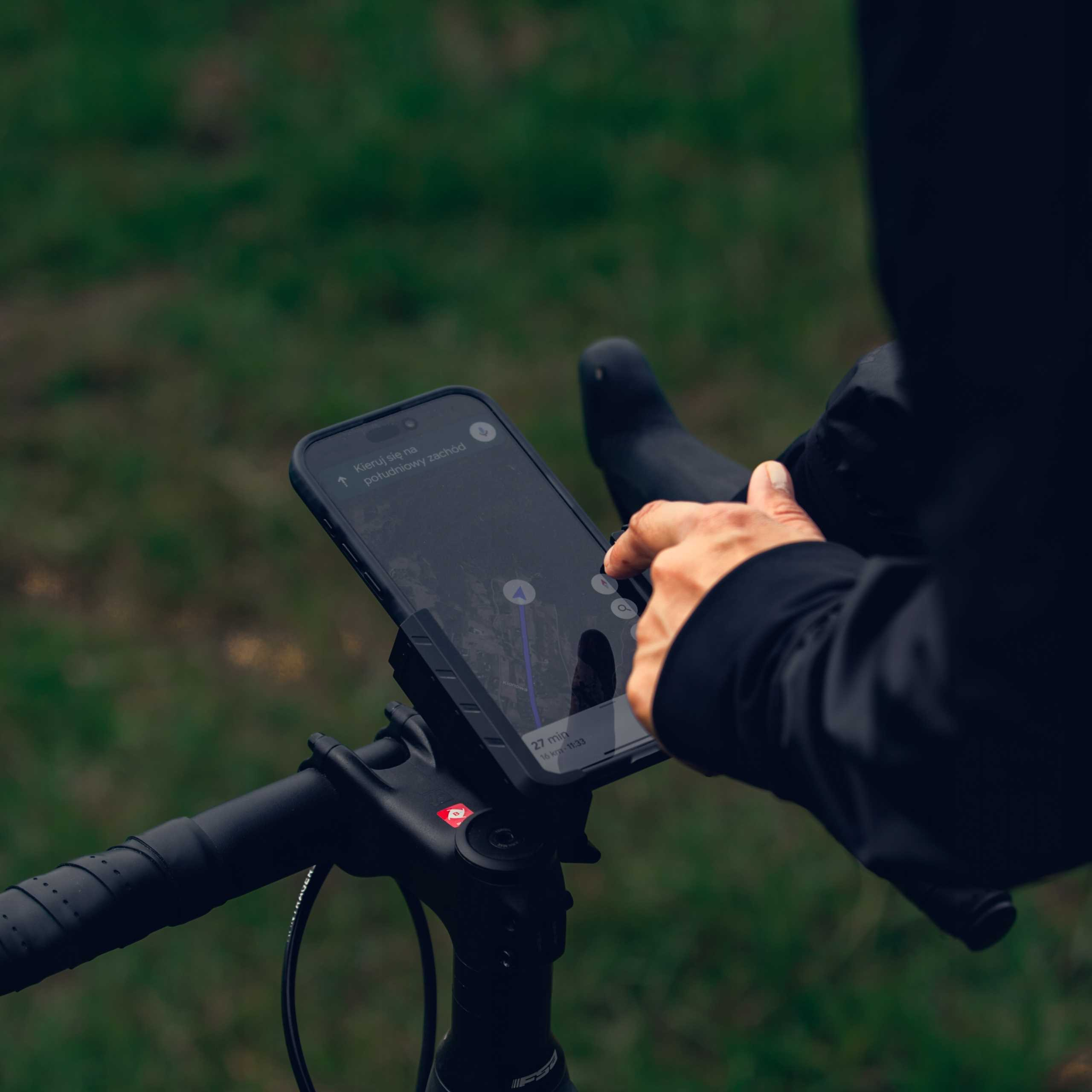 Dłoń obsługuje smartfon z aplikacją nawigacyjną na kierownicy roweru.