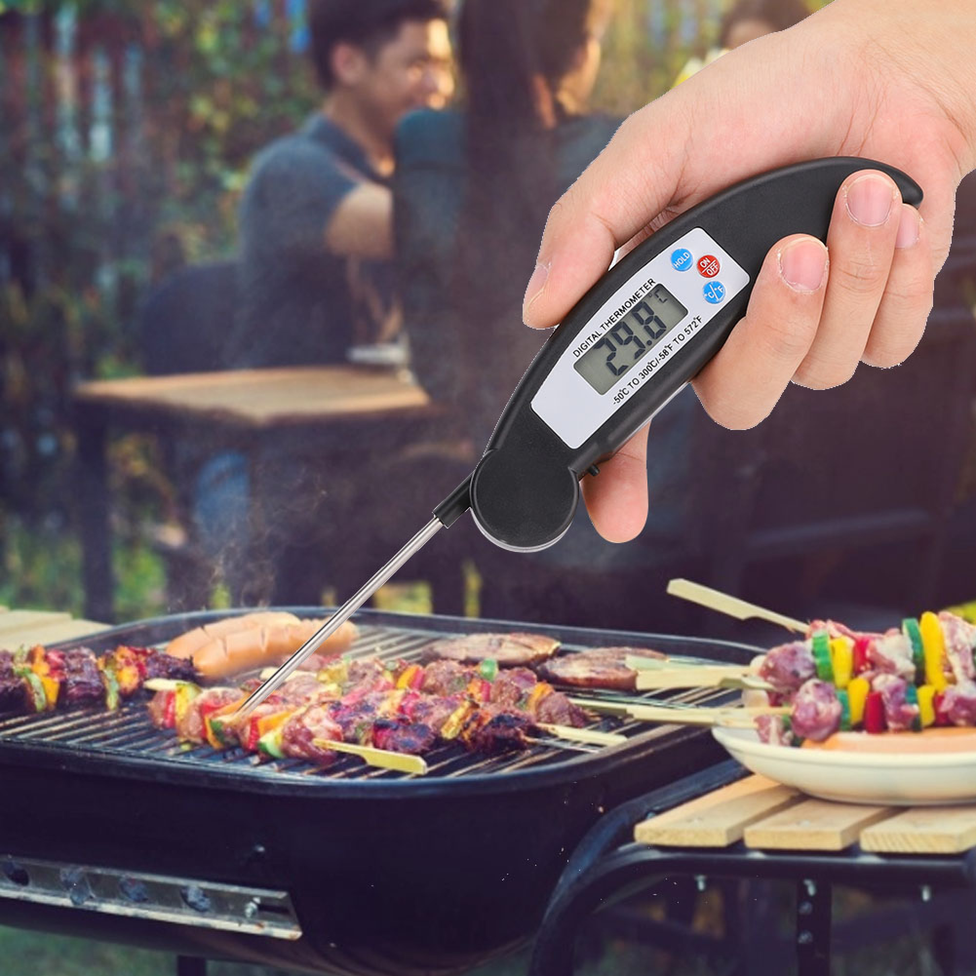 Eine Hand hält ein digitales Thermometer über einem Grill mit Fleischspießen und Würstchen.