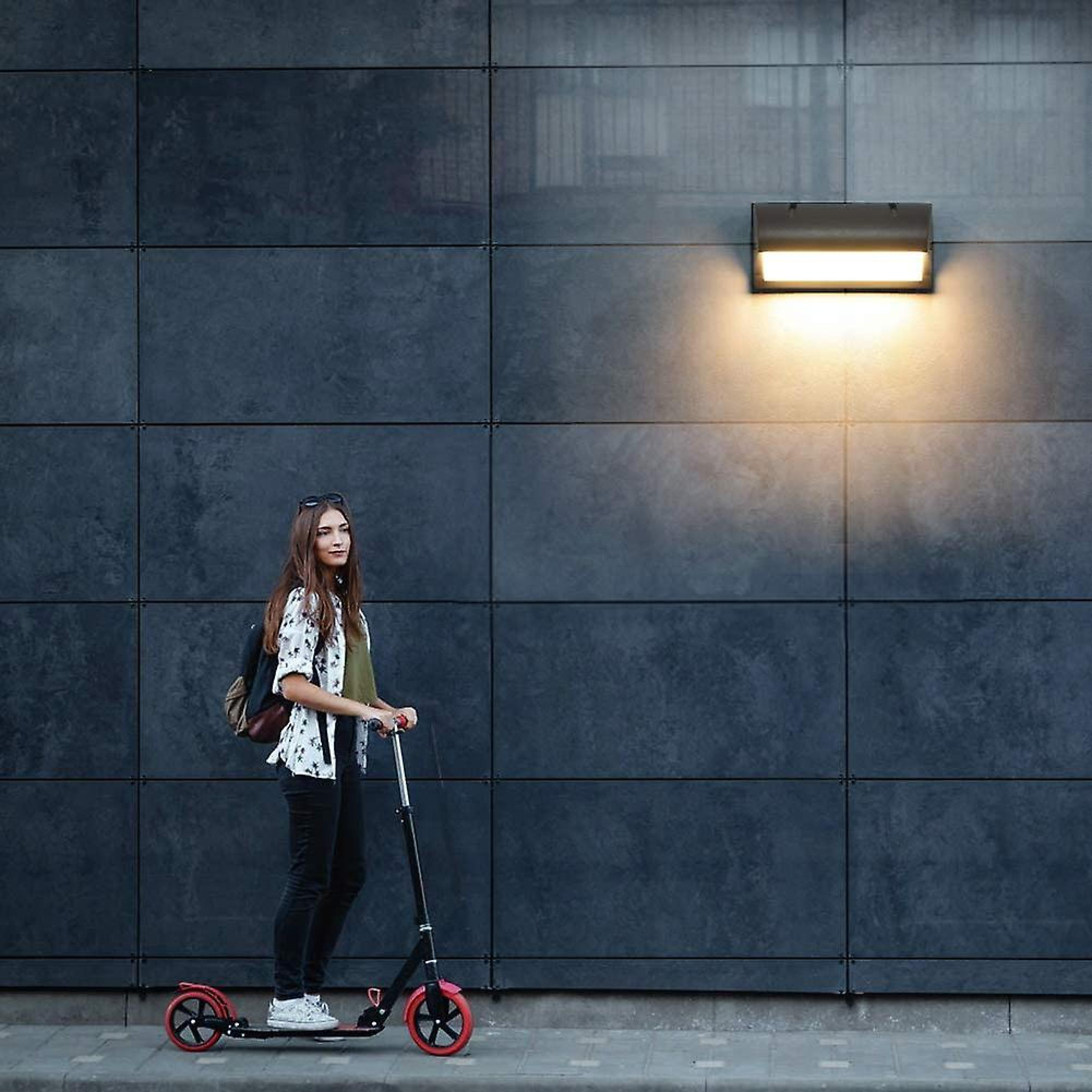 Eine Frau mit Rucksack steht auf einem Roller vor einer dunklen Wand.