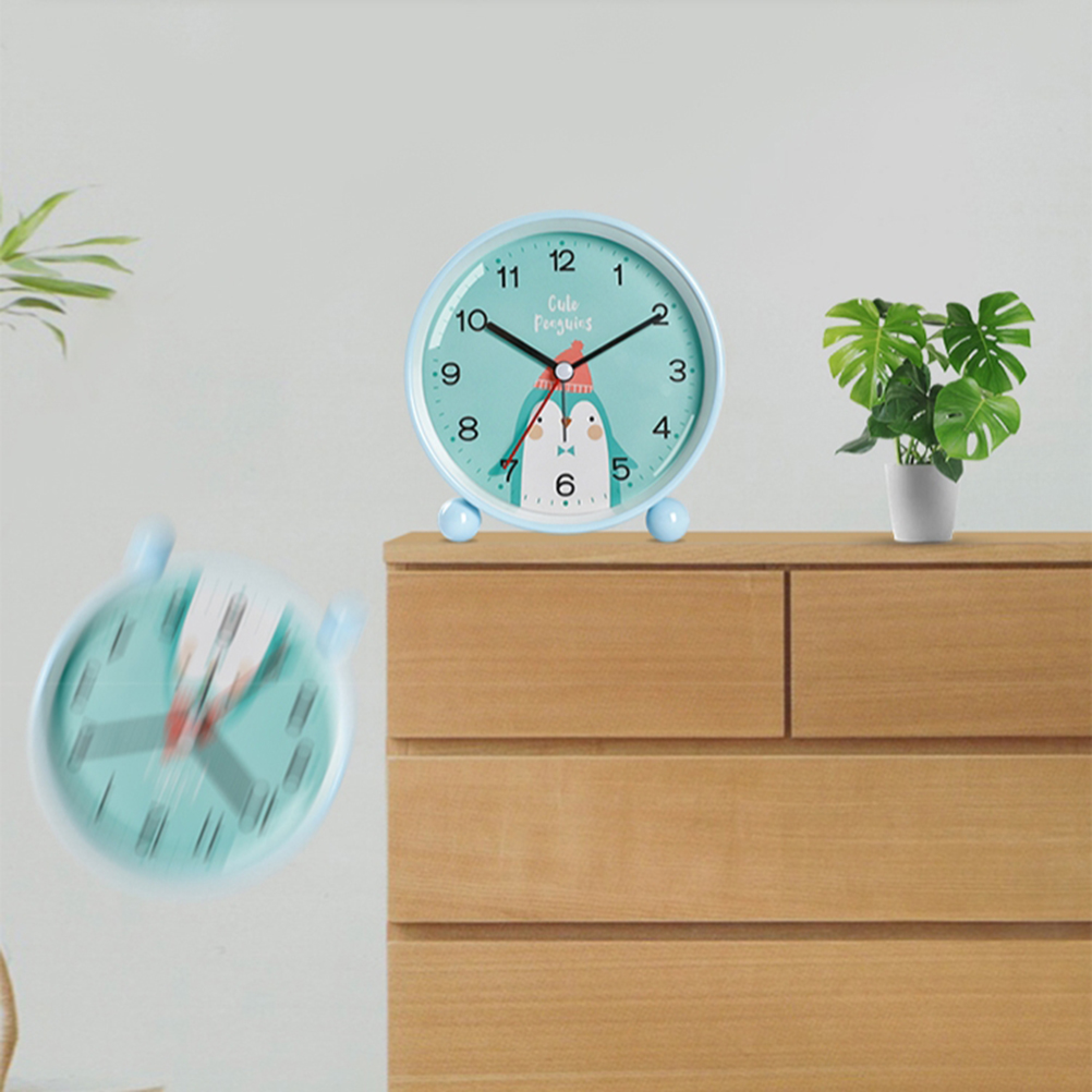 Ein blauer Wecker mit Pinguin-Motiv steht auf einer Holzkommode neben einer grünen Pflanze.