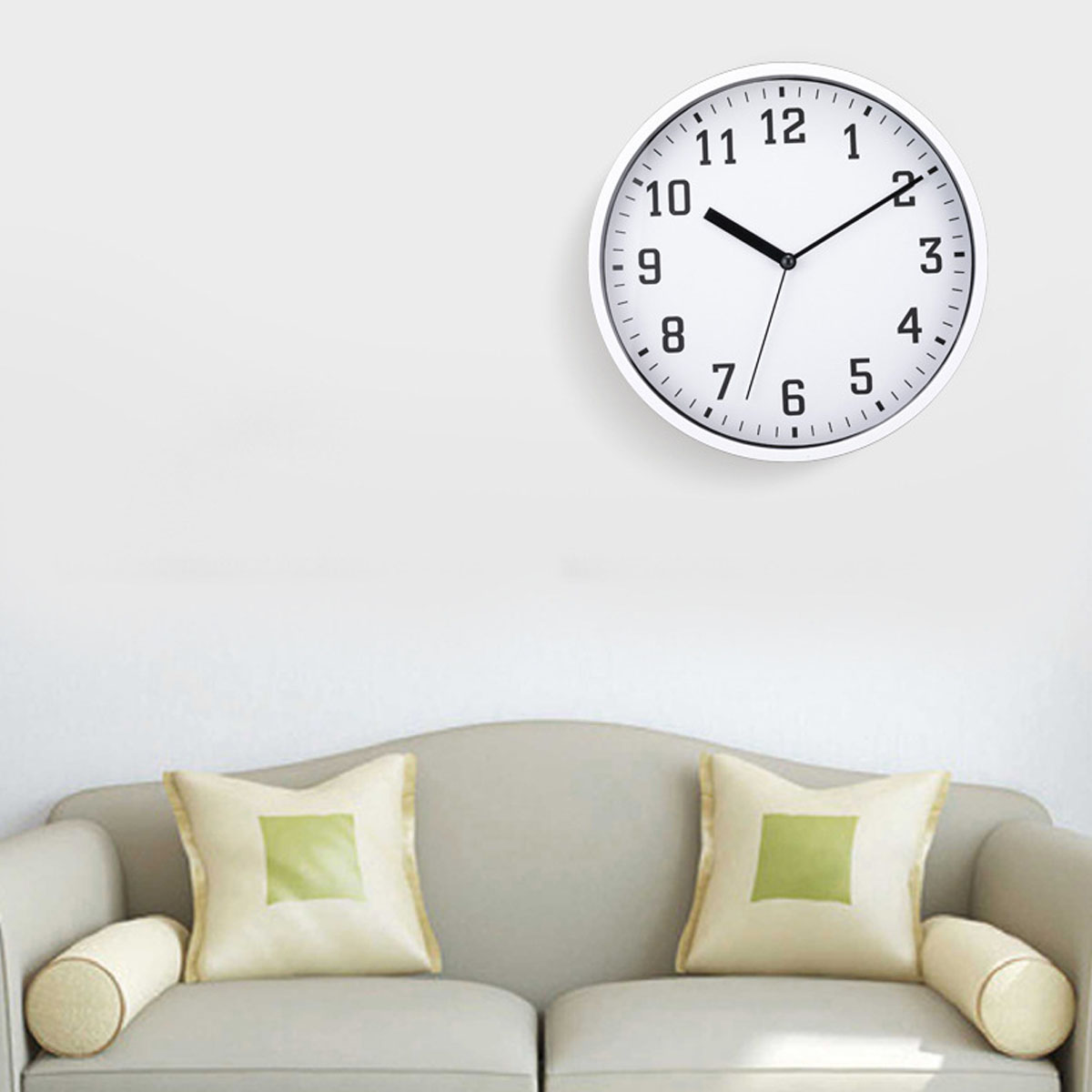 Eine weiße Wanduhr mit schwarzen Ziffern hängt über einem hellgrauen Sofa mit Kissen.