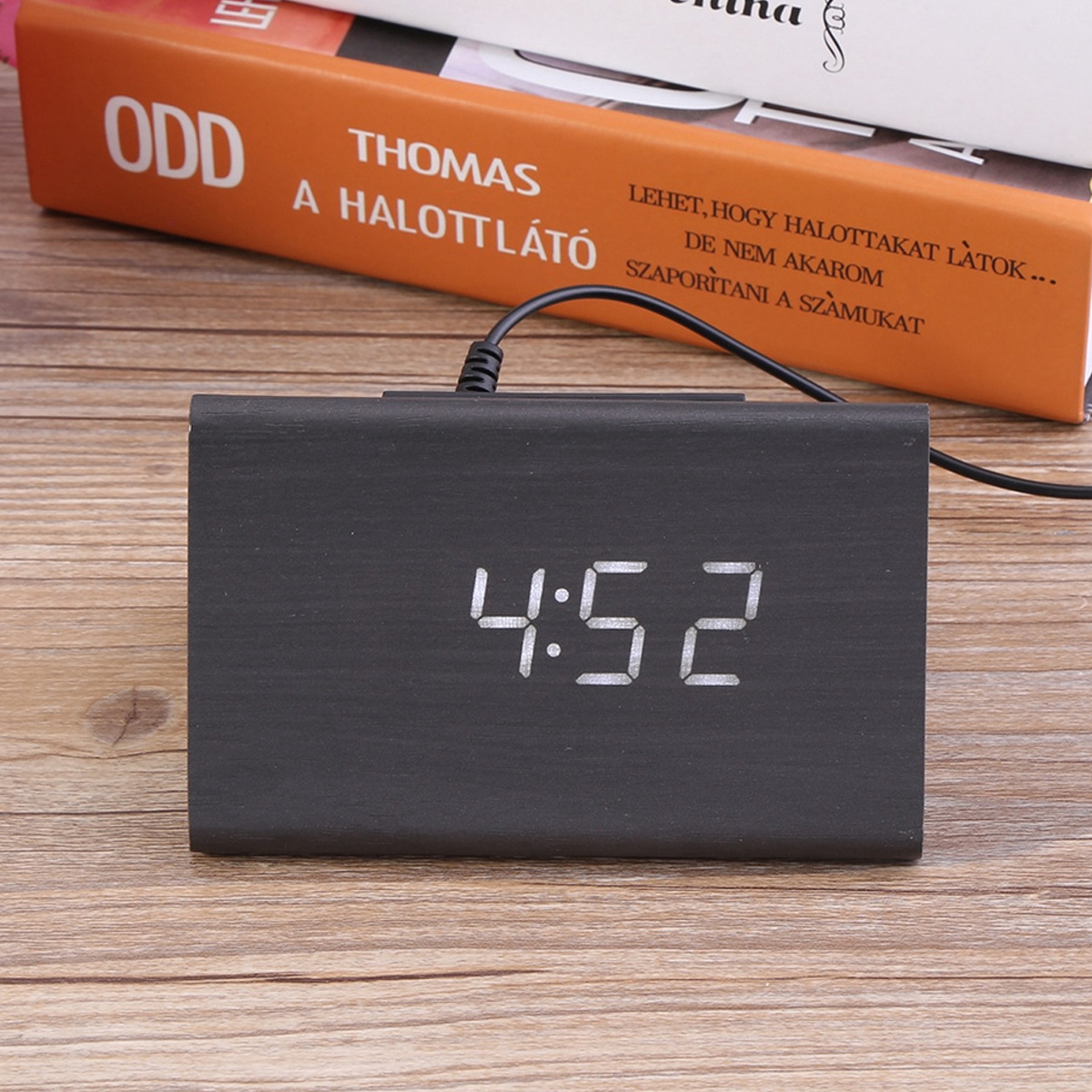 Ein schwarzer Digitalwecker zeigt 4:52 auf einem Holztisch mit Büchern im Hintergrund.